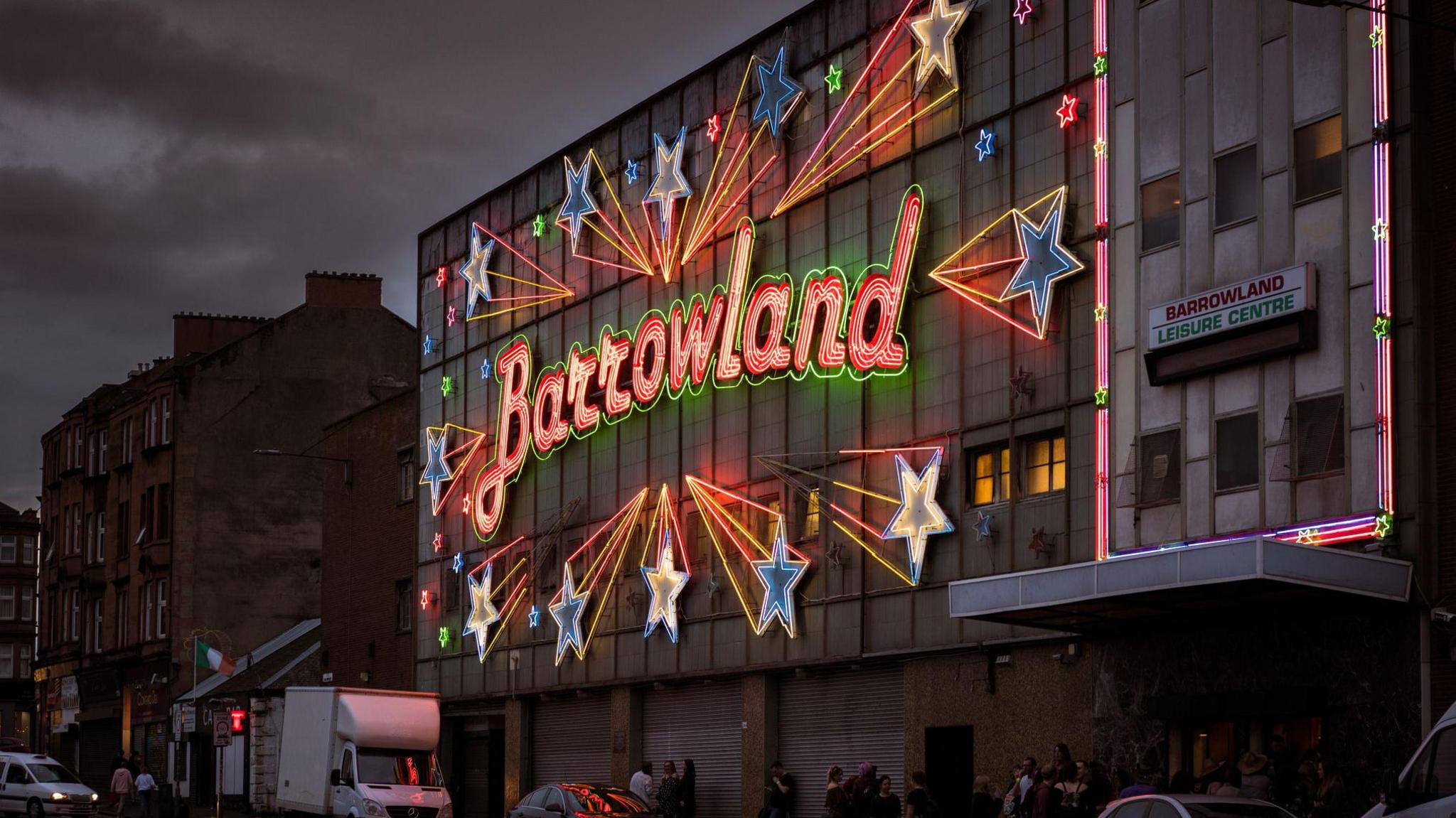The neon sign on the front of the Barrowland ballroom in Glasgow is lit up on a dark night. The word "Barrowland" is in red letters, surrounded by blue and yellow stars