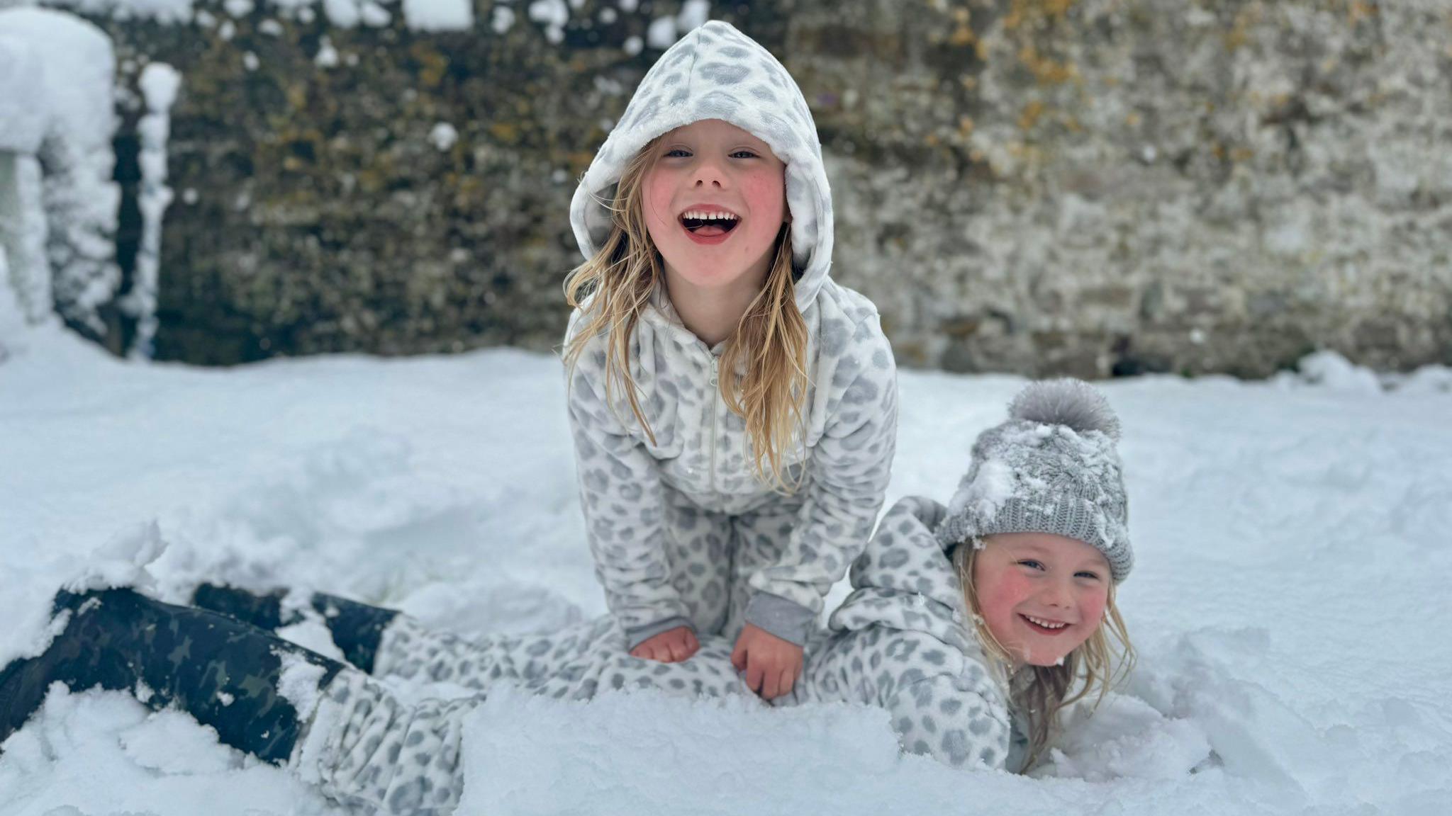 Two girls playing in snow in a field. They both have blonde hair and are about 6 years old. They are wearing white and grey animal print onesies with hoods, snow boots, and one of them is wearing a grey hat.