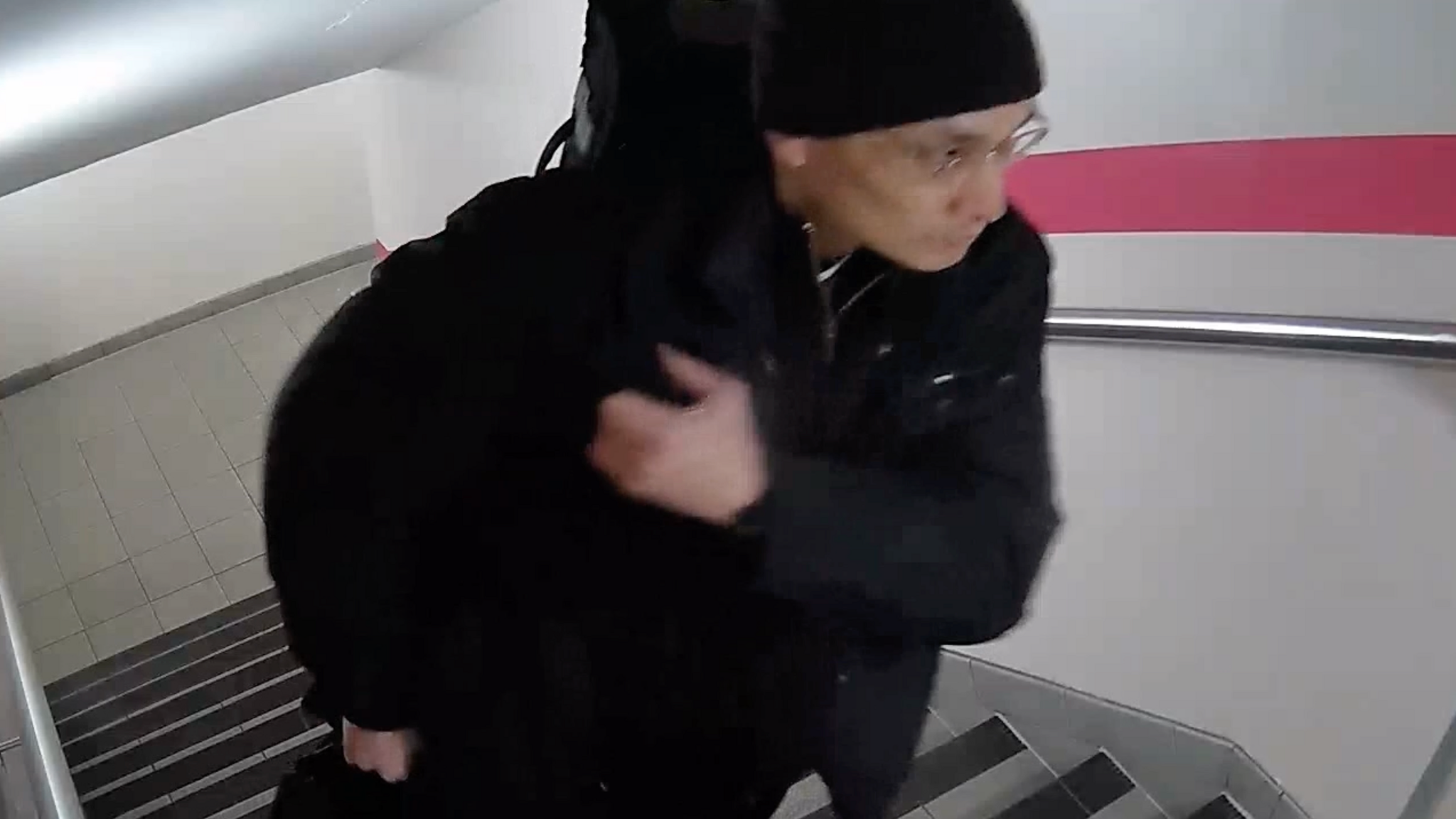 Still from CCTV footage of a man all in black wearing a black hat and carrying a bag walking up stairs.