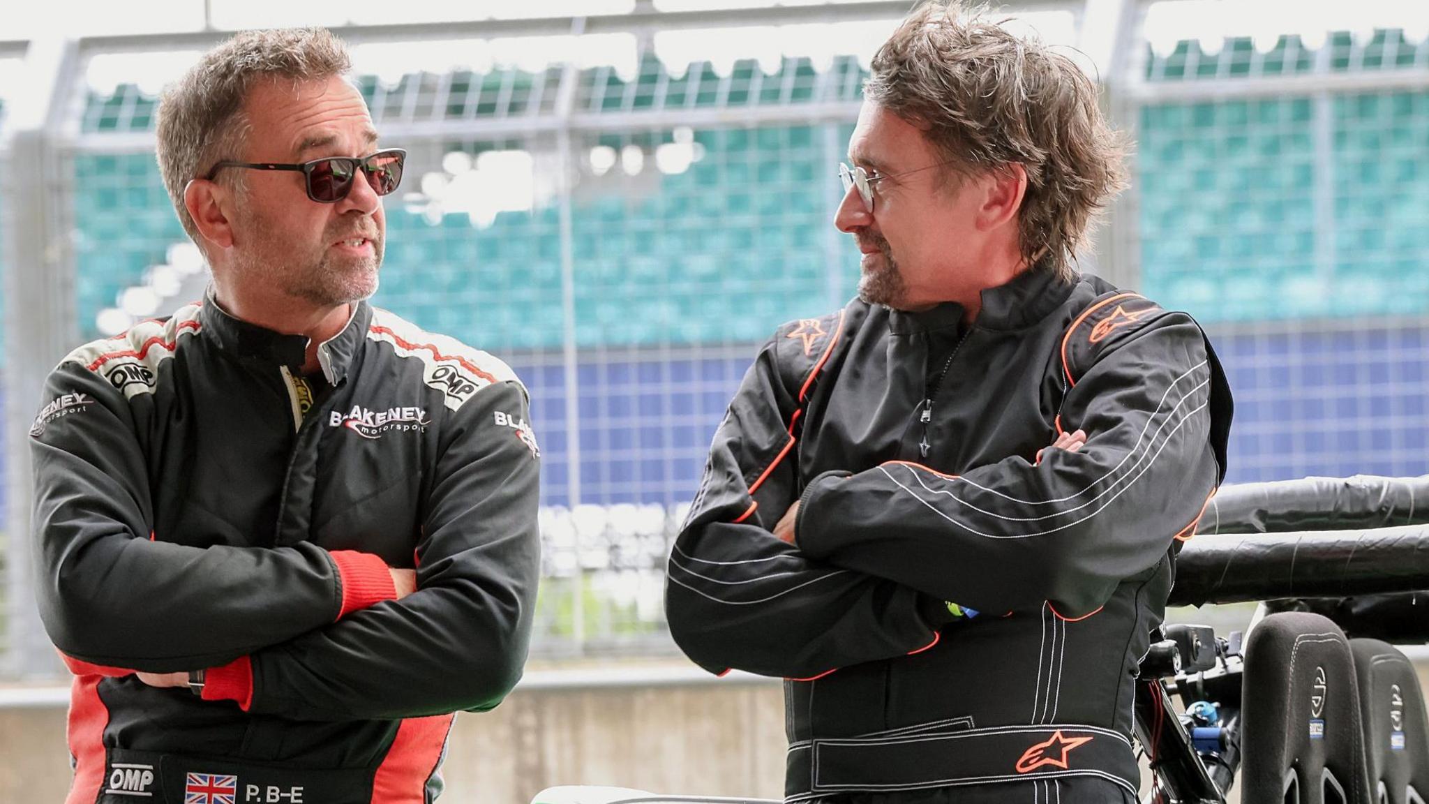 Two men in black motor racing suits with their arms folded talking to one another. Behind them is a fence and a grandstand