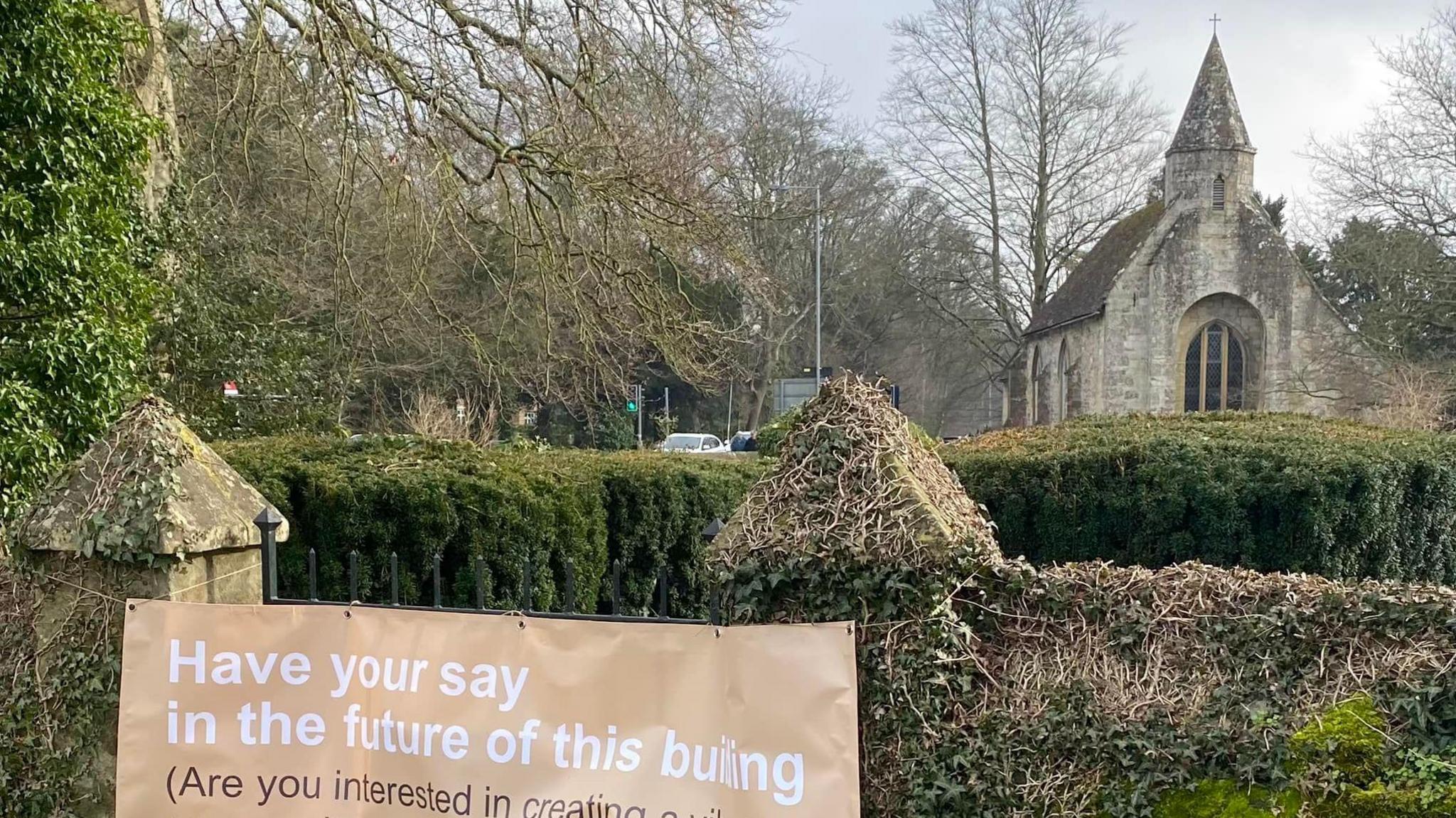 A stone church surrounded by a yew hedge with a sign reading "Have your say in the future of this building".