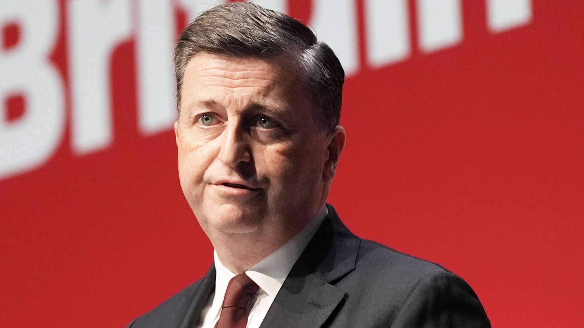 Head shot of Douglas Alexander on a stage delivering a speech against a red backdrop. He has black hair, combed in a side shed and is wearing a grey suit, white shirt and maroon tie.