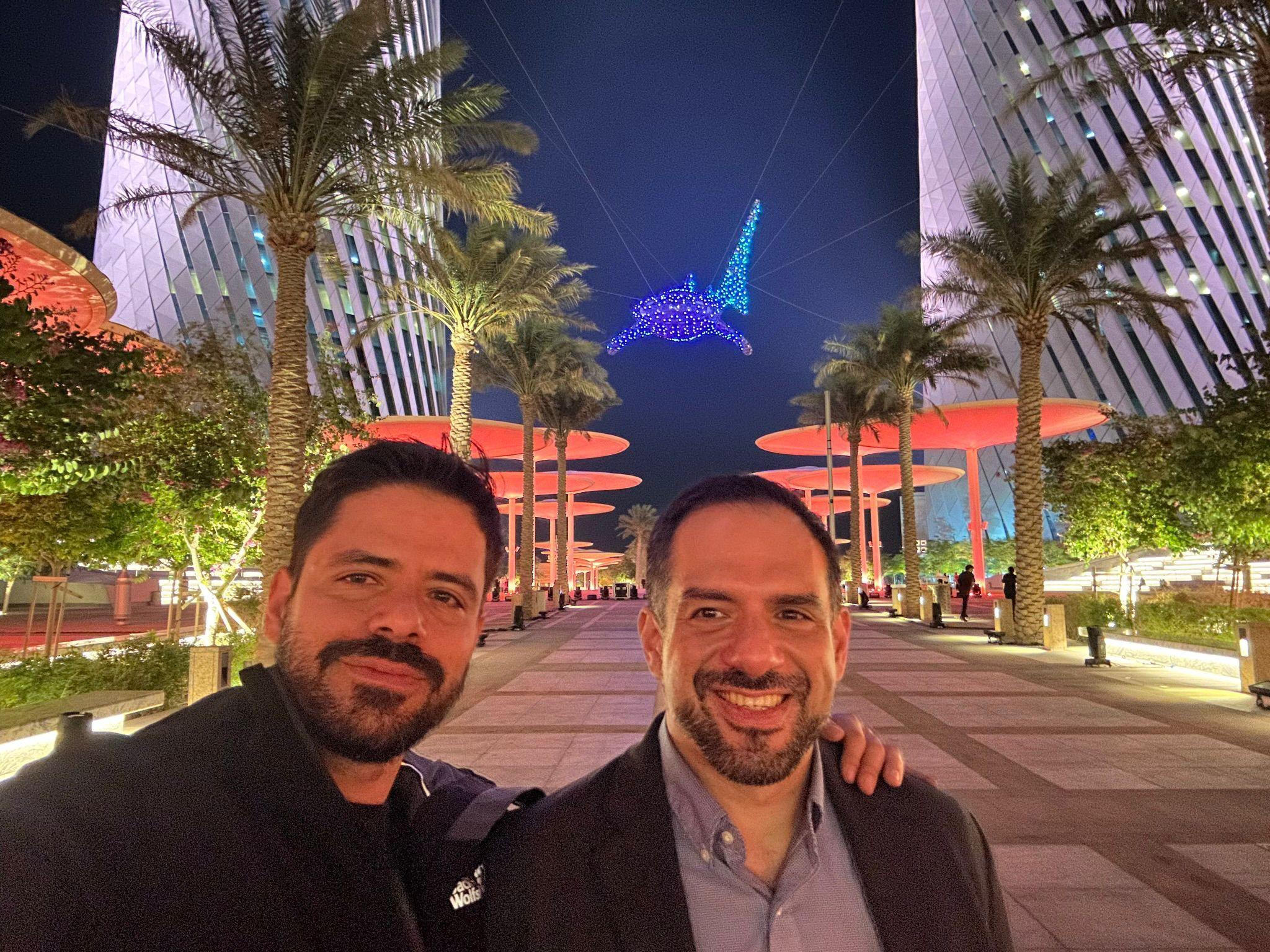 Two brothers pose in front of a lit-up decoration shaped like a shark suspended in the sky. They are both wearing jackets and both have black hair with beards.