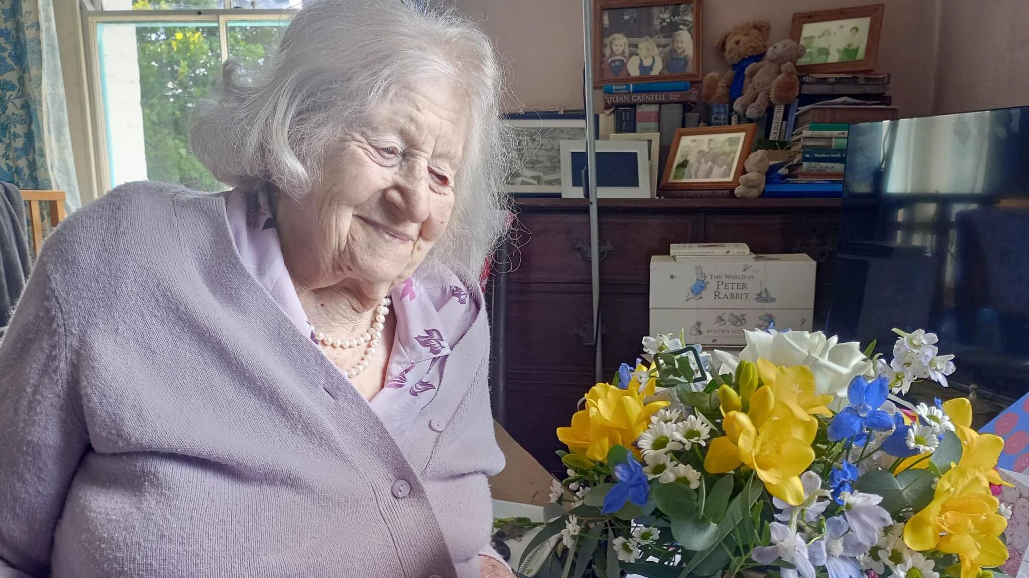 Anne Baker wears a lavender cardigan and shirt as she looks down at a bouquet of flowers with yellow, blue and white.