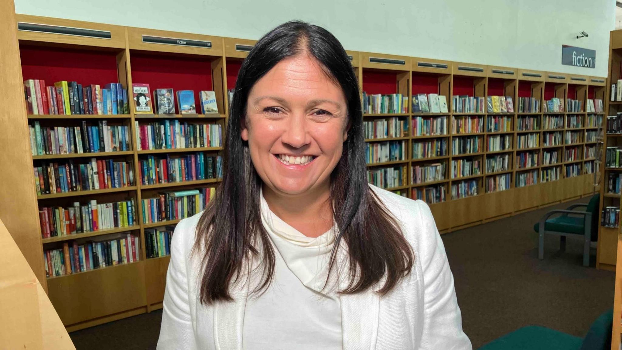 Lisa Nandy, a Labour MP, is standing in a library with shelves of books behind her. She has long dark hair and is smiling widely. She is wearing a white suit jacket and white top.