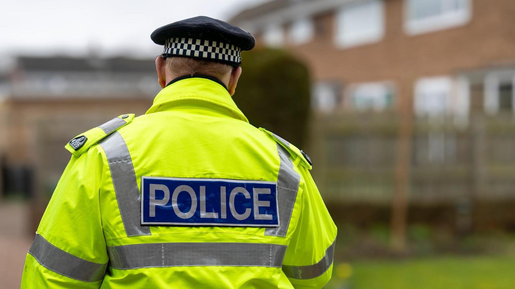 A generic image of the rear of a police officer wearing a black hat with a white and black checked band, and a yellow hi-vis jacket.