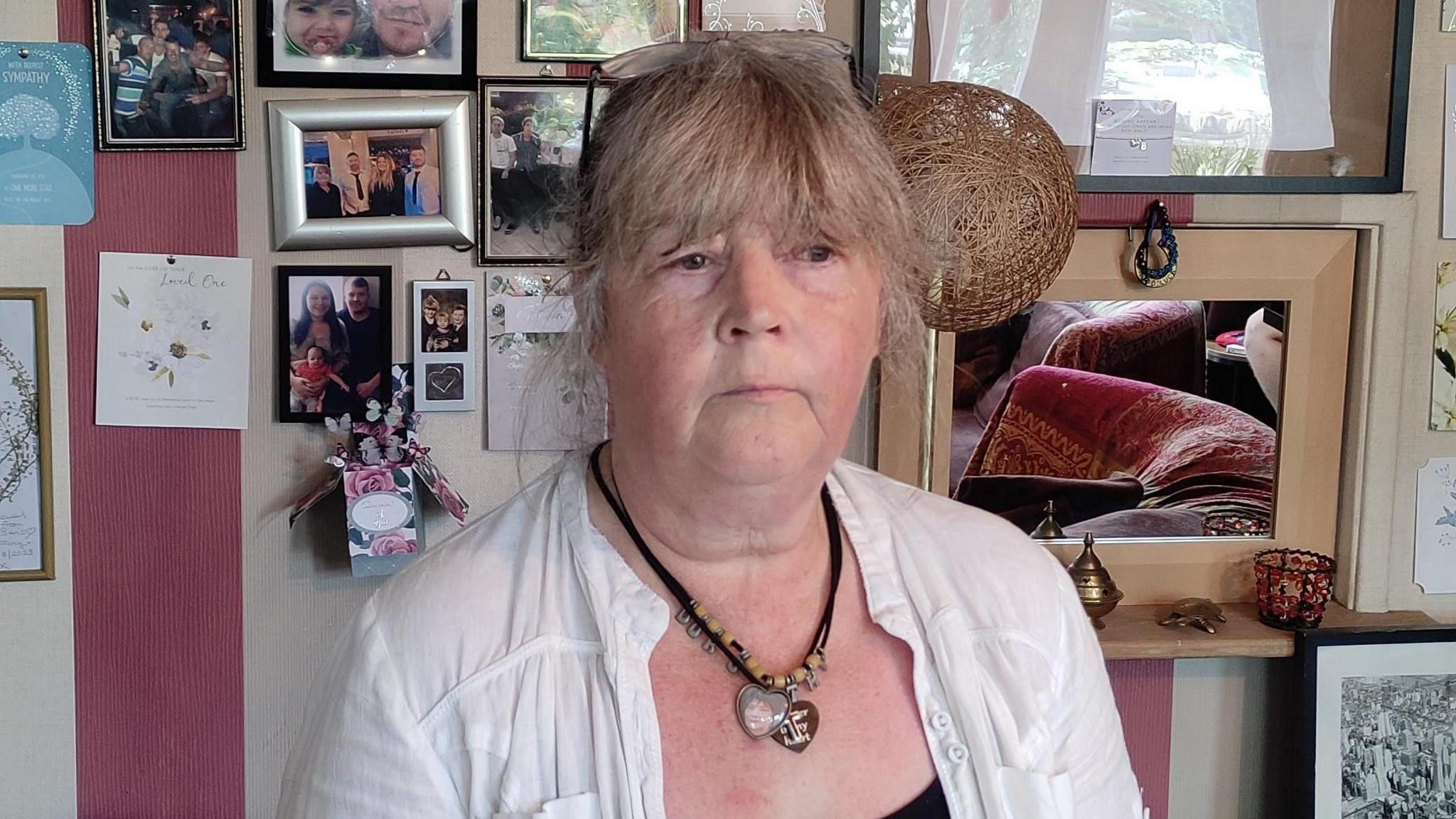 Dot is wearing a white shirt and two necklaces. She has glasses sitting on her head and is standing in front of a red and white striped wall filled with photographs. 