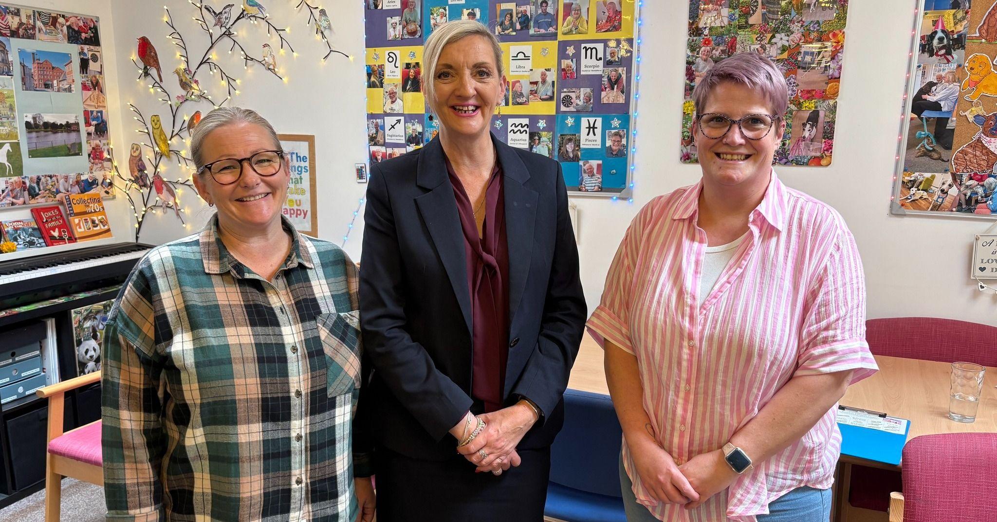 Three women smile at the camera. The woman to the left is wearing a green and white checked shirt and glasses. Beside her is a taller woman with blonde hair that is tied back. She is wearing a navy suit with a burgundy pussy-bow shirt beneath. The woman on the right of the image is wearing a pink and white striped shirt and she has short purple hair and glasses. They are standing in a room which is decorated with pictures and posters.