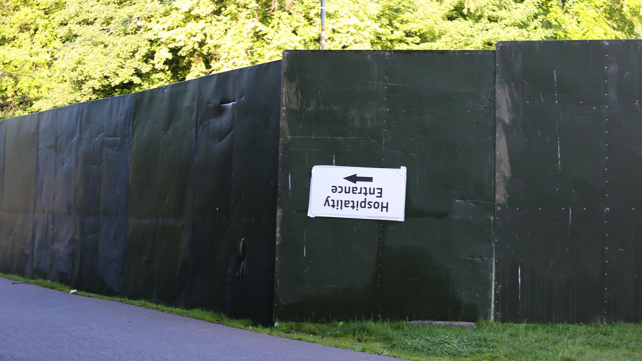 A hospitality entrance sign hangs upside down on a green metal fence in front of foliage in Heaton Park
