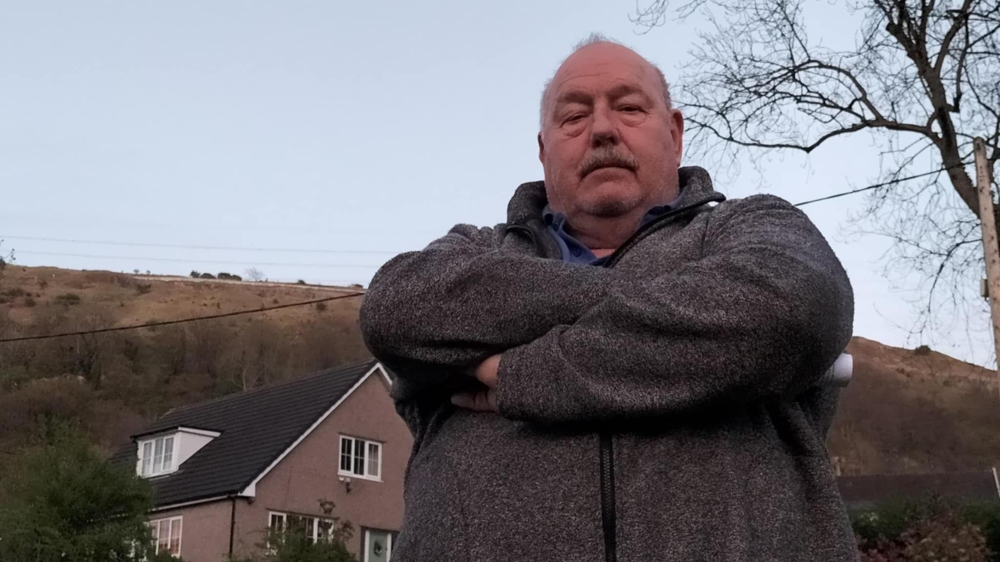 A bald man with a moustache wearing a grey fleece, looking angry with his arms crossed across his body. Behind him are houses and hills.