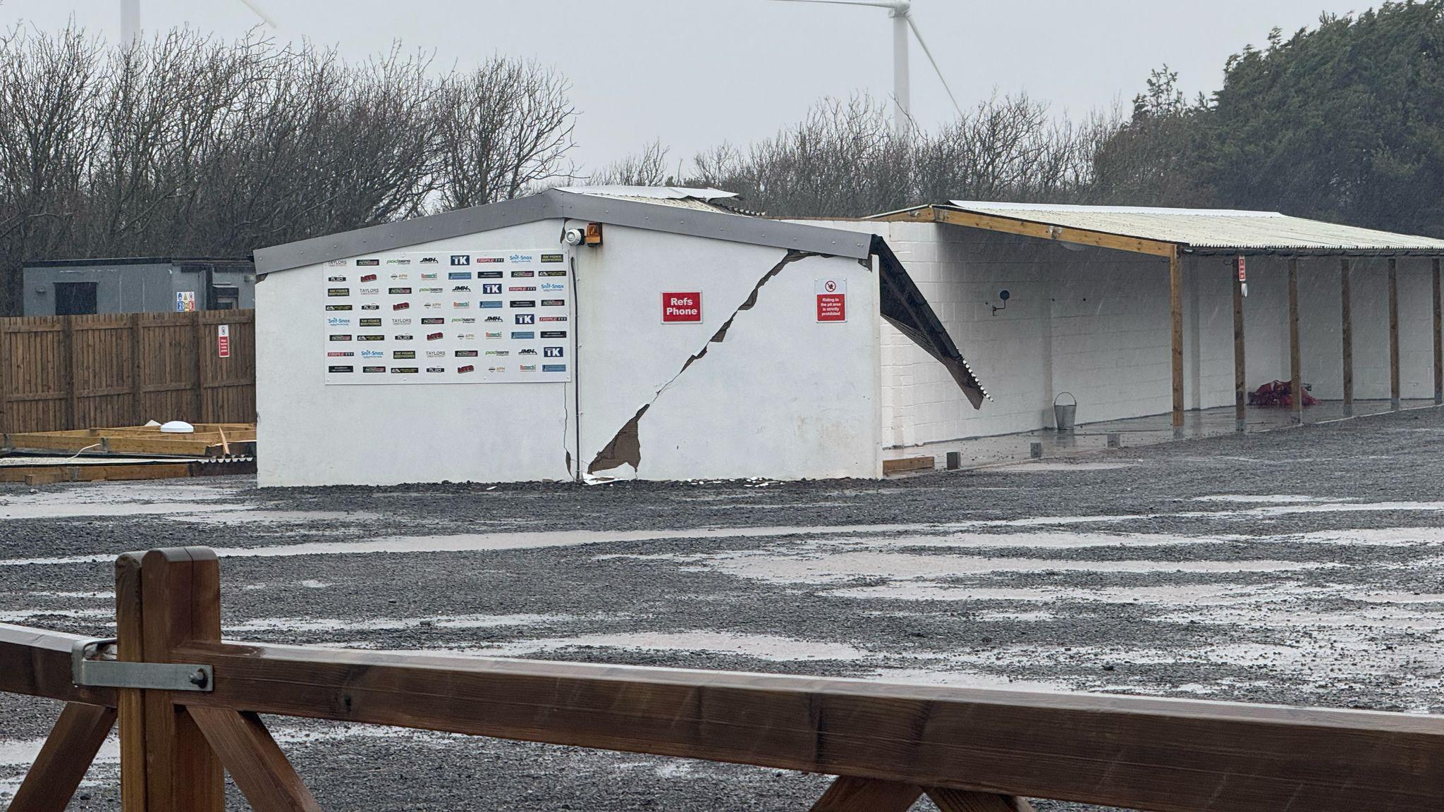 The side of the Workington Comets pits building. The white gable wall has a large diagonal crack in it and its roof is hanging off. 