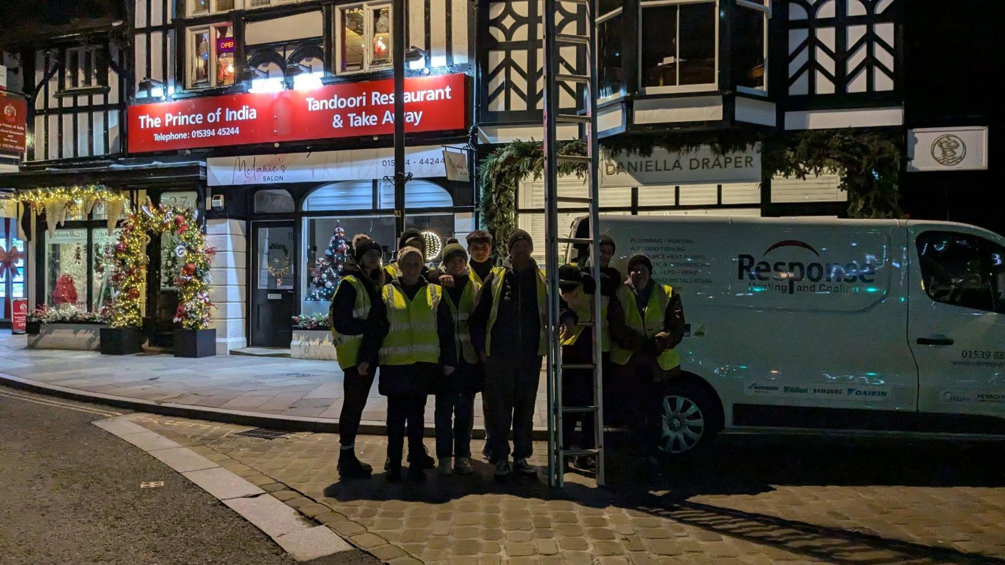 Eight volunteers wearing high-vis vests in Windermere town centre. They are posing for the shot outside an Indian restaurant, holding a tall set of ladders.