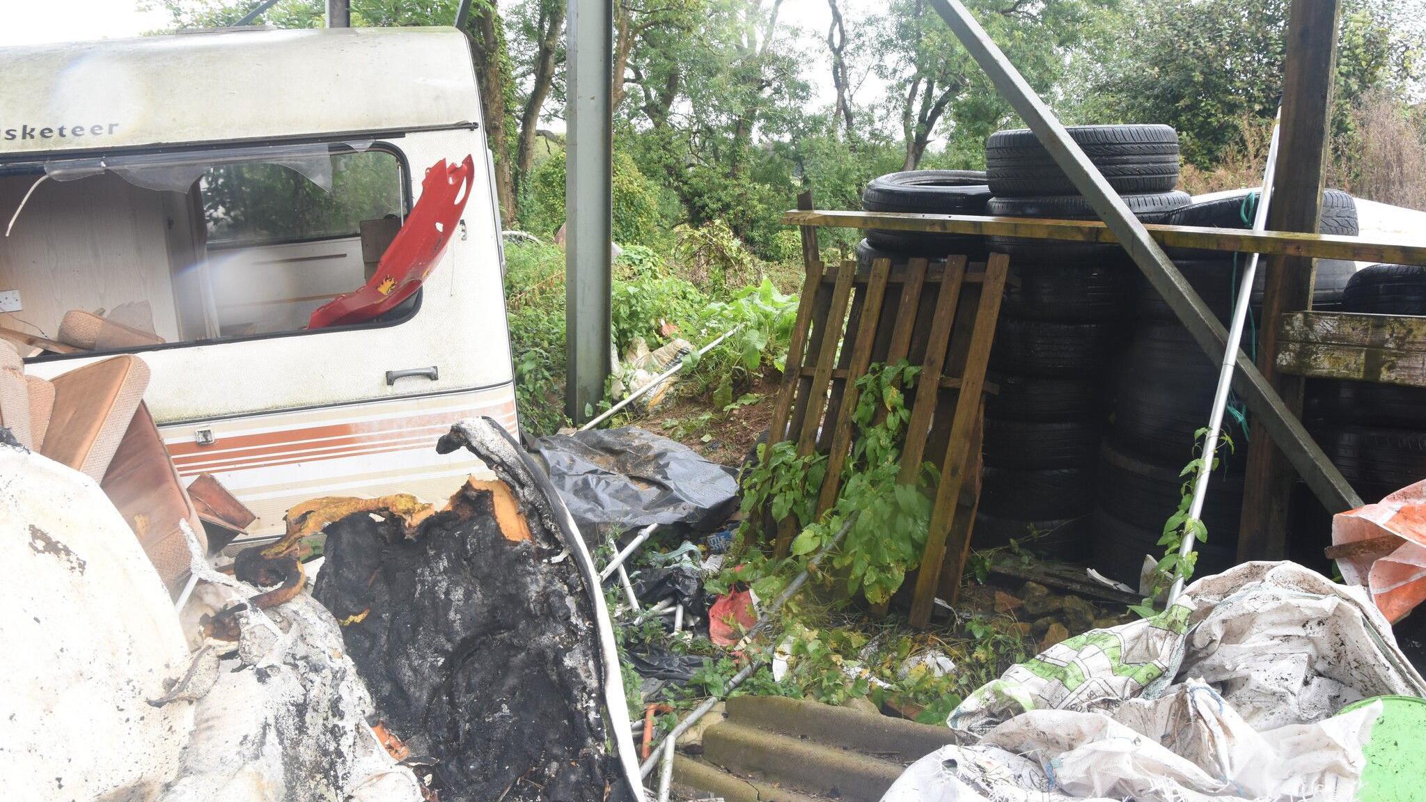 An abandoned caravan inside a vacant lot. The windows are smashed and there is debris scattered around- including stacked tyres, wooden crates, burnt leather furniture, a corrugated tin roof and an old duvet. The whole site is overgrown and surrounded by trees.
