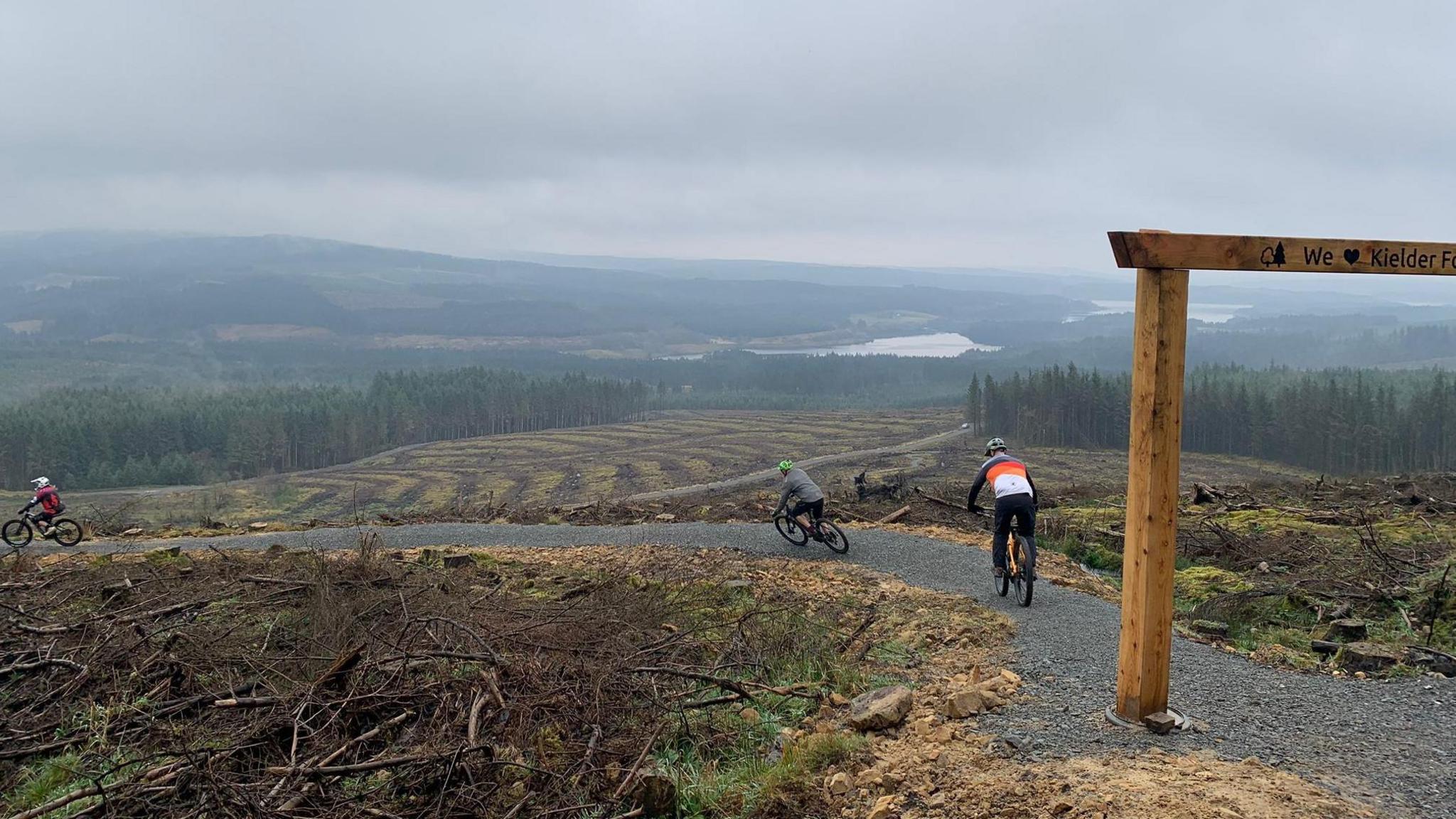 Two cyclists set off down a trail path with Kielder Forest in the distance. 