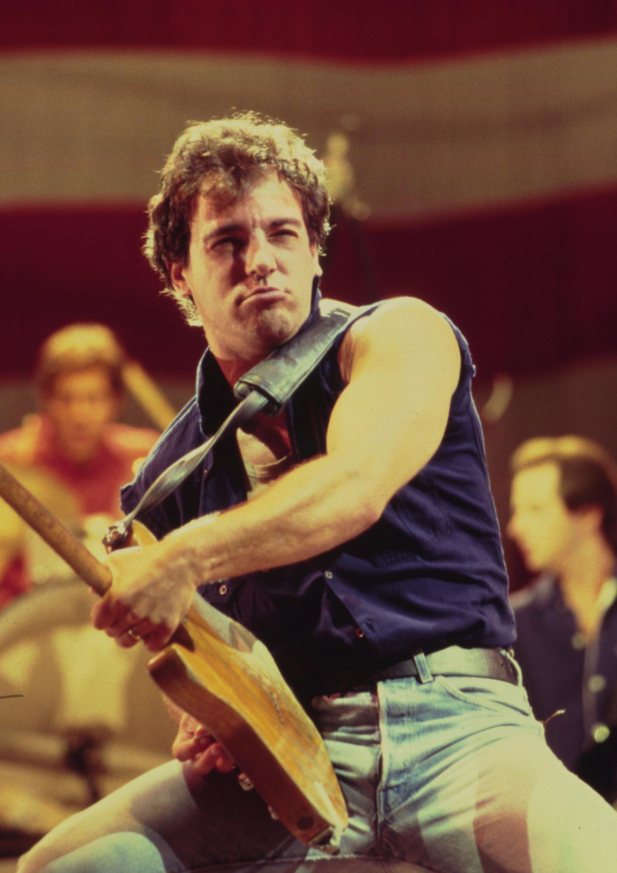 Bruce Springsteen on stage in the 1980s, he's wearing a blue vest, light blue jeans and is holding his guitar out far in front of him.