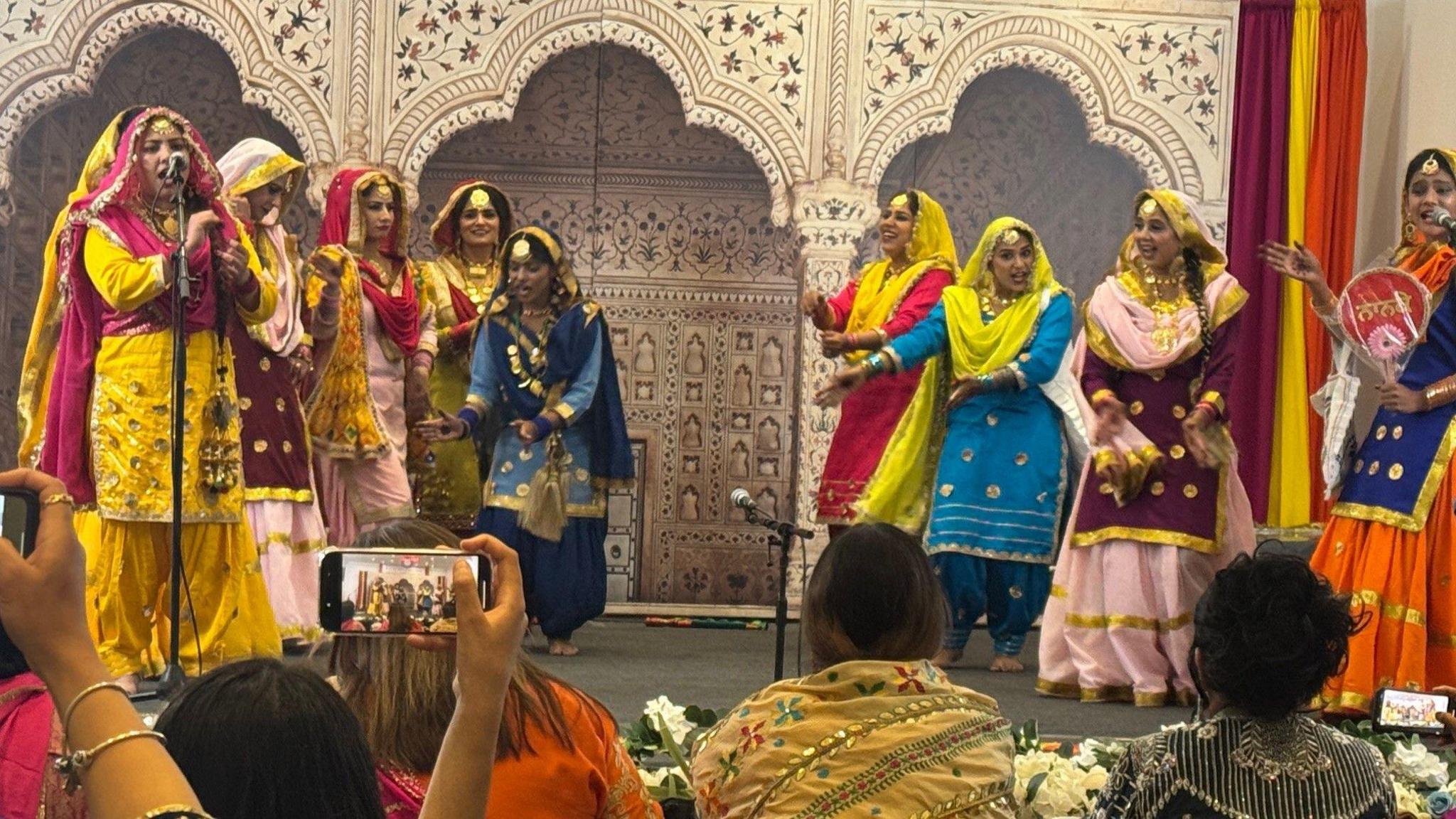 Nine women with dark hair dancing on a stage in traditional Indian dresses made of gold, yellow, pink, red and blue fabric with decorative arches on the wall behind them.