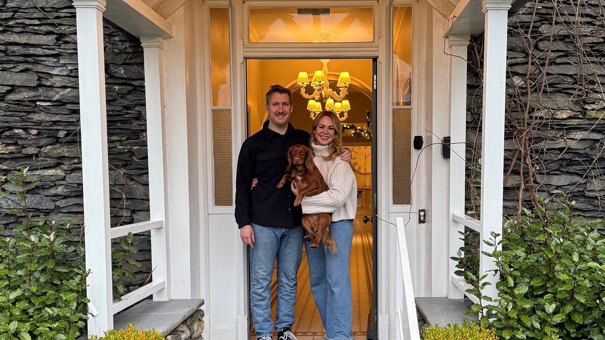 Lauren and Greg Foggo standing in front of the hotel door holding their dog, a ginger spaniel.