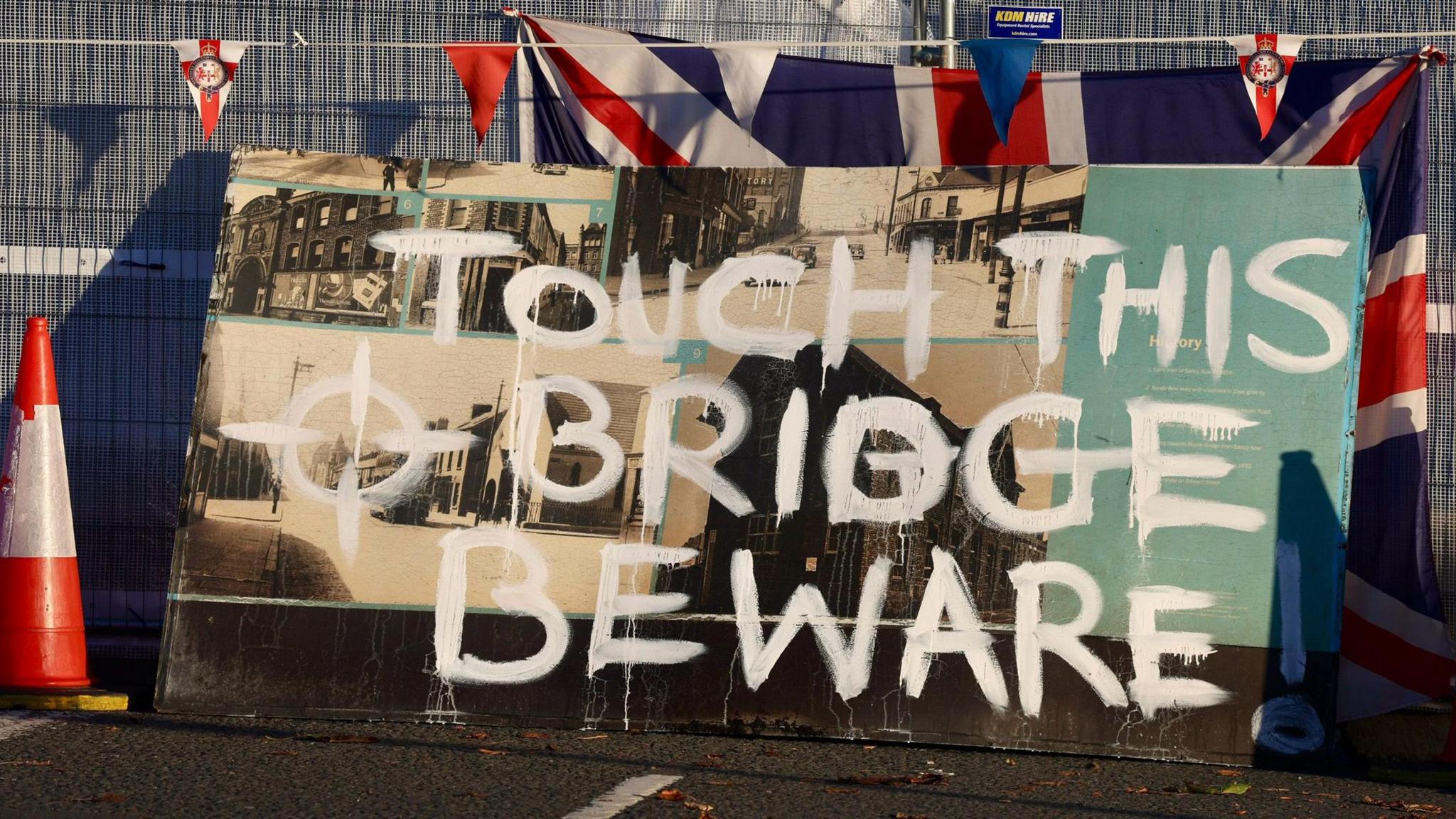 A fence covered in red white and blue union jacks with a sign saying "touch this bridge, beware! Traffic cones are dotted along it. 