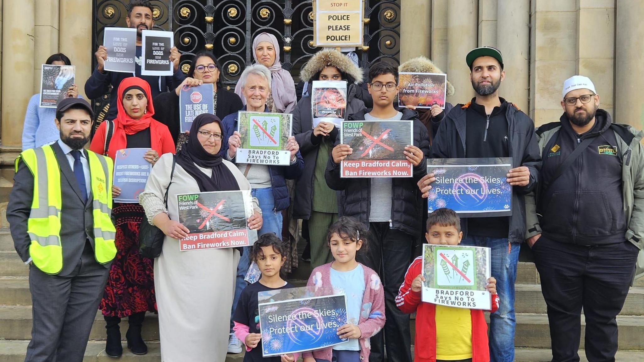 A group of people, a mix of adults and children, stand on steps holding up signs with messages such as "Ban Fireworks Now" and "Silence the Skies, Protect Our Lives".