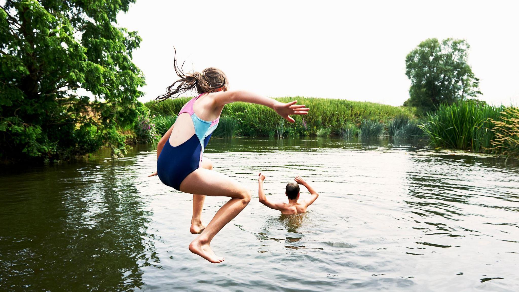 A boy and a girl jumping into a lake surrounded by trees and reeds