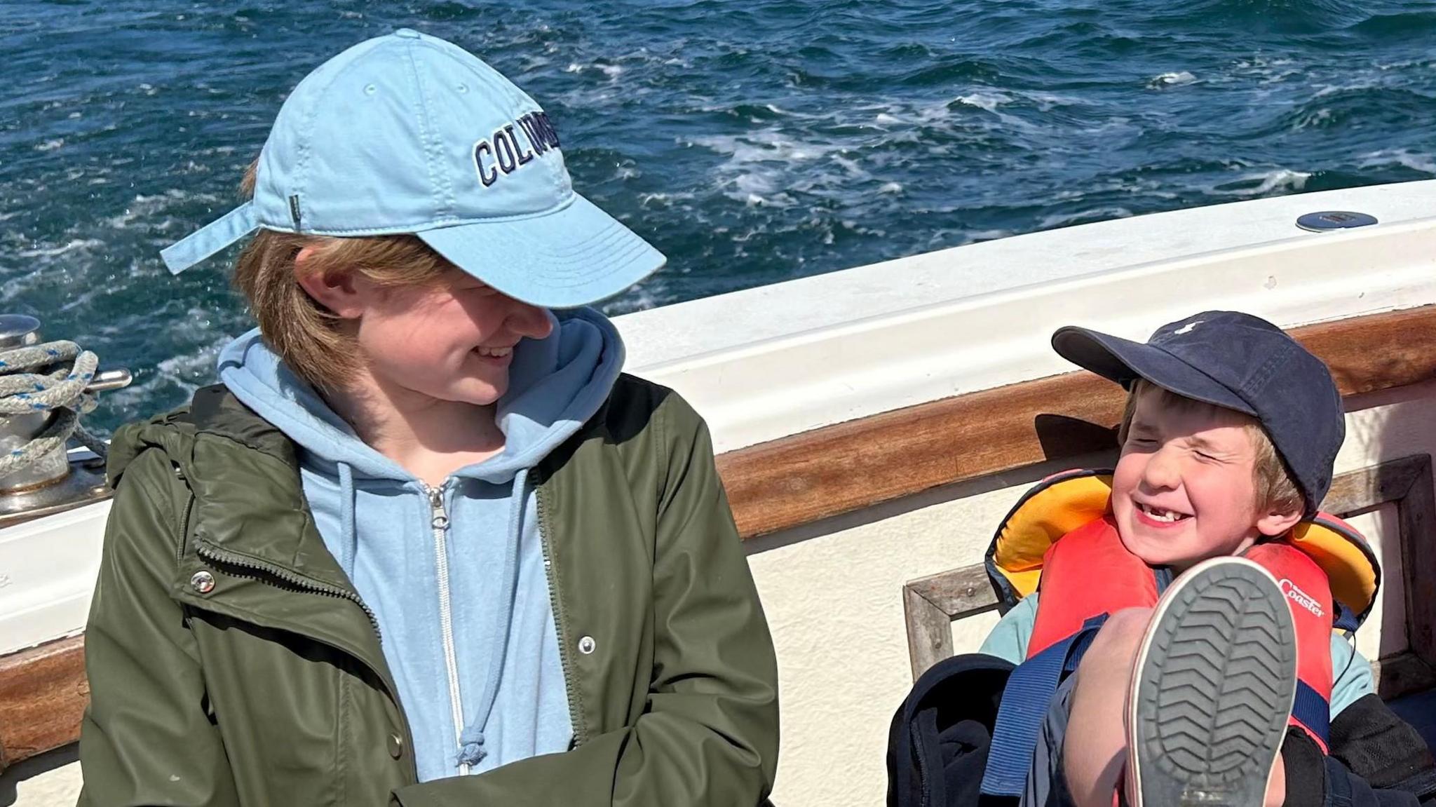 Alex is wearing a dark green coat with a light blue hoodie and a light blue cap. Teddy is smiling for the camera and is wearing a lifejacket and a blue cap. He has his foot up, facing the camera. They are both on a boat, with water behind them both.