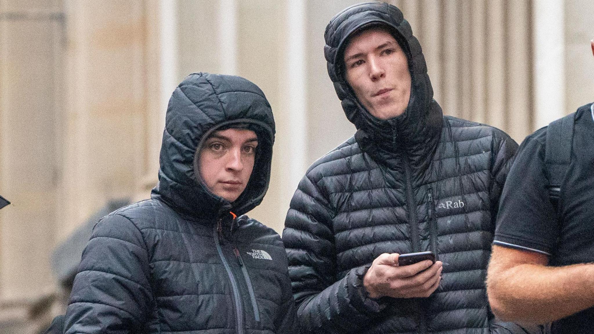 Two men in black coats, stood with their hoods up outside a court building. One of them is holding a mobile phone.