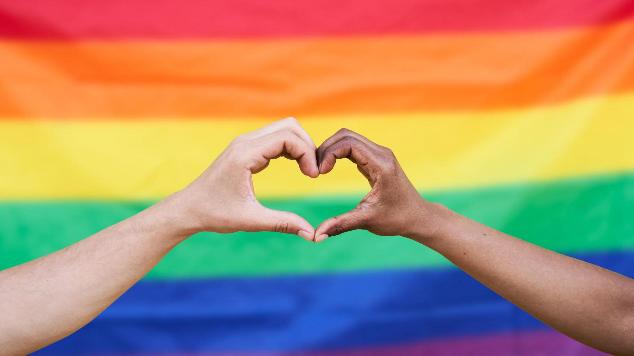 Two hands form a love heart against a rainbow flag in the background. 