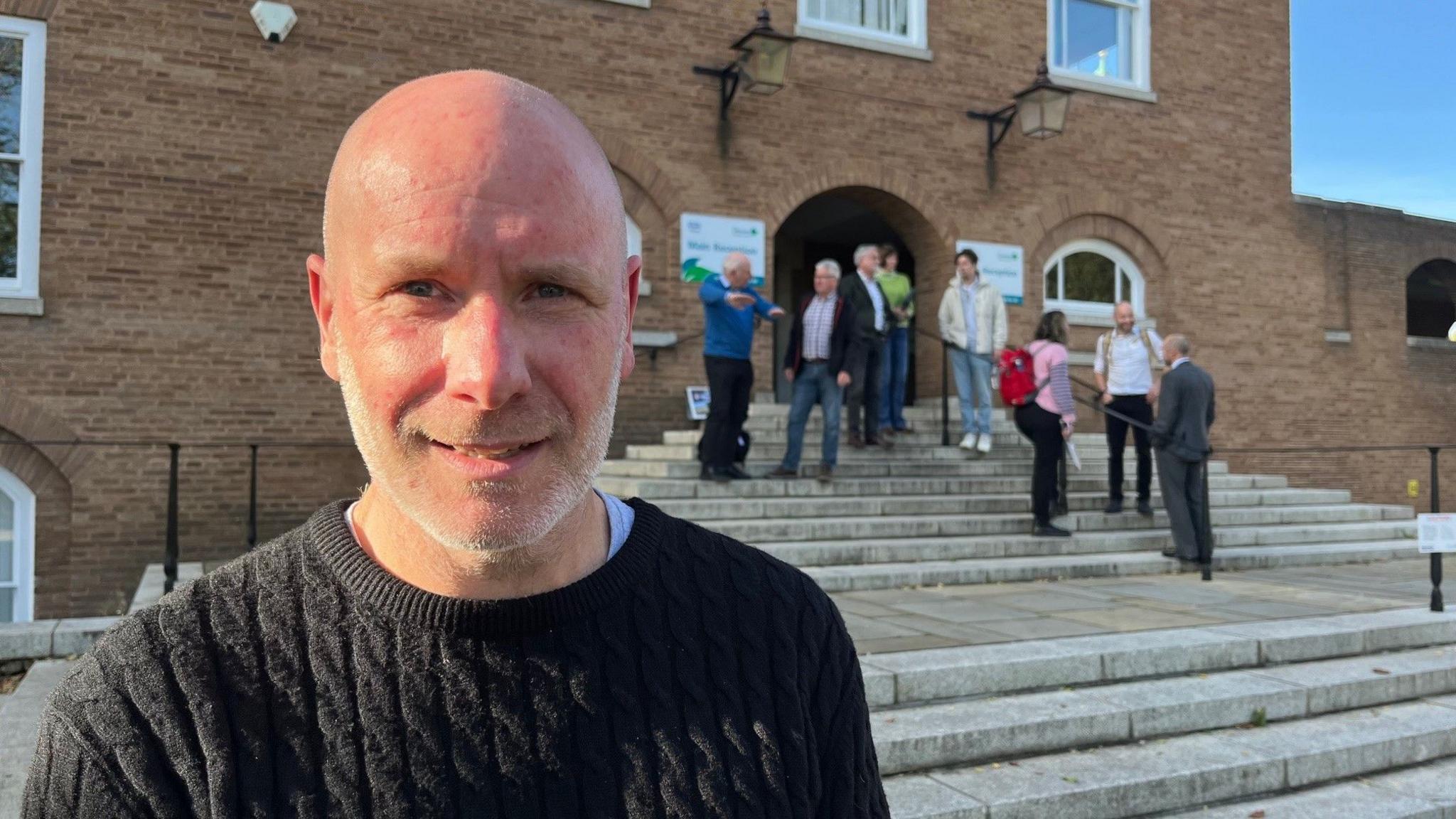 Steven Badcott in a blue ribbed jumper in front of steps at County Hall with people in the background