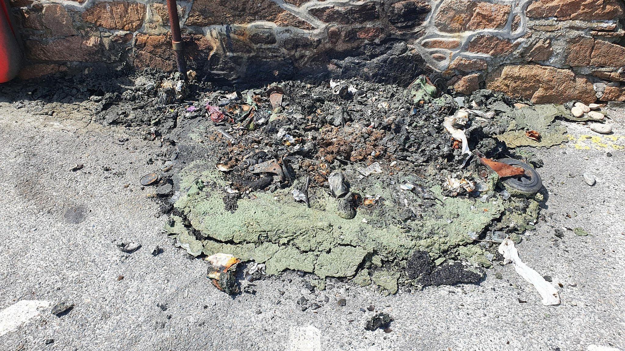 The remnants of a melted plastic bin on the floor in front of a wall. The remnants include melted plastic, ash and rubble.