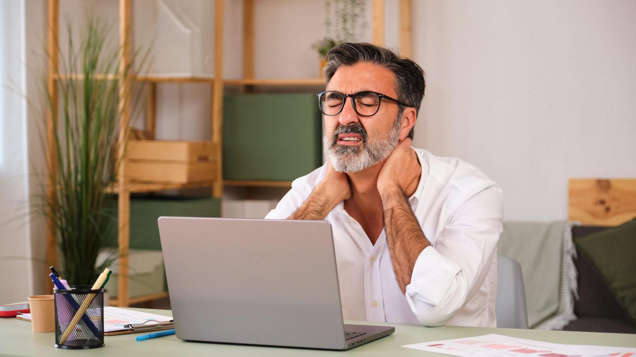 A man with a laptop on his lap is hunched over and rubbing his neck. He is in pain and is likely working on the computer.