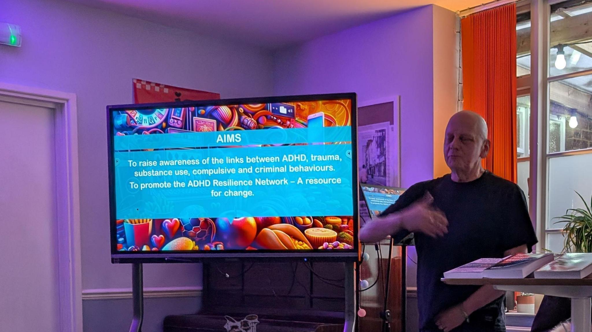 Colin McGregor-Paterson talking next to an electronic screen. The screen is showing text to highlight some of the aims of the cafe. The text reads: "AIMS: To raise awareness of the links between ADHD, trauma, substance use, compulsive and criminal behaviours. To promote the ADHD Resilience Network - a resource for change."