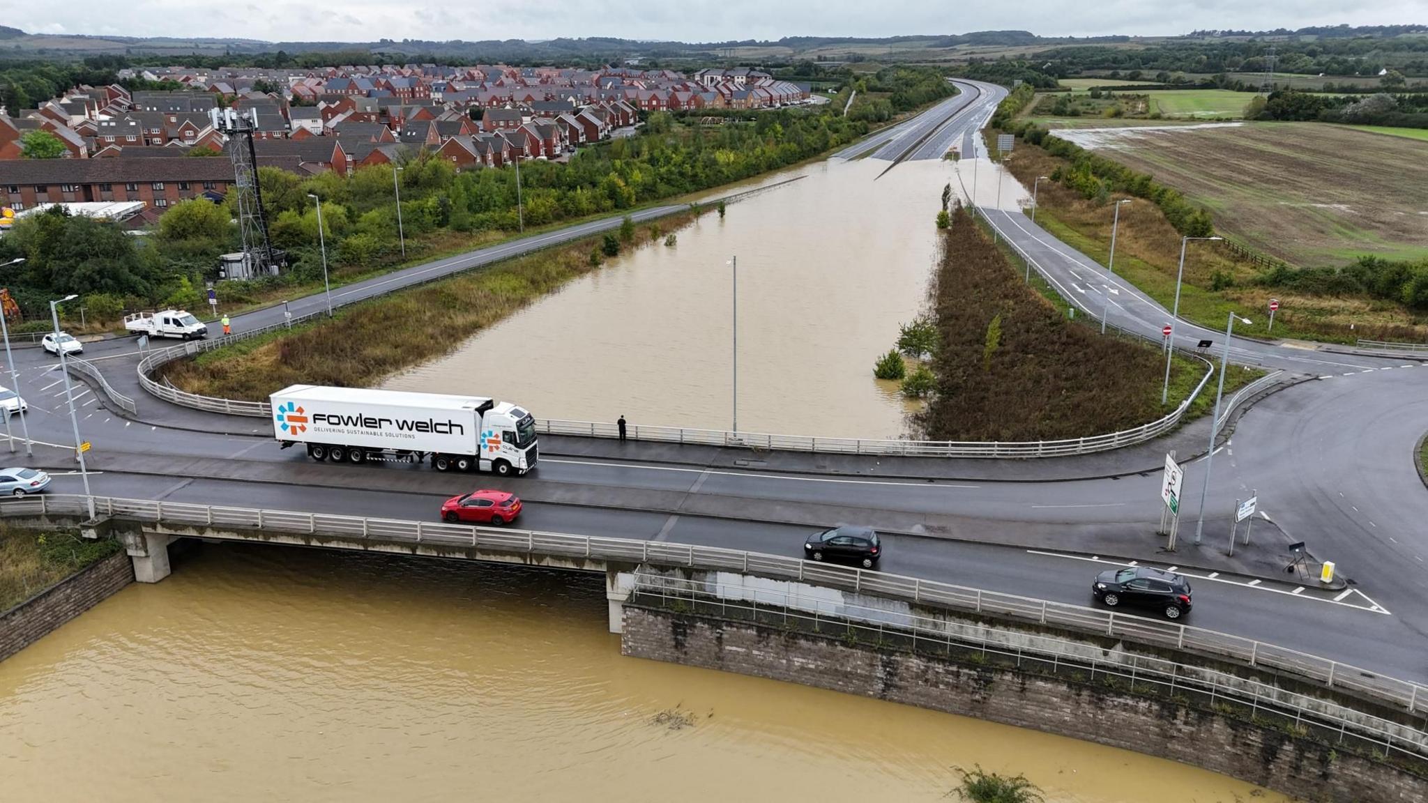 Aerial photograph of traffic crossing the bridge over the flooded A421 at Marston Moretaine in Bedfordshire. The dip under the bridge is several hundred metres long and the water is a murky light brown colour. 