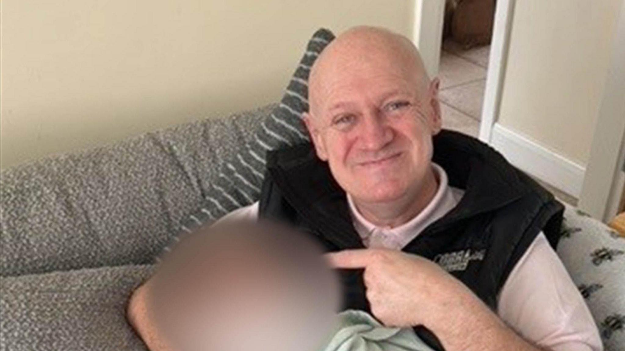 Christopher Hanley smiles pointing to a blurred out image of another person while sat on a grey sofa. He is bald and wearing a pink polo shirt covered by a black gillet. 