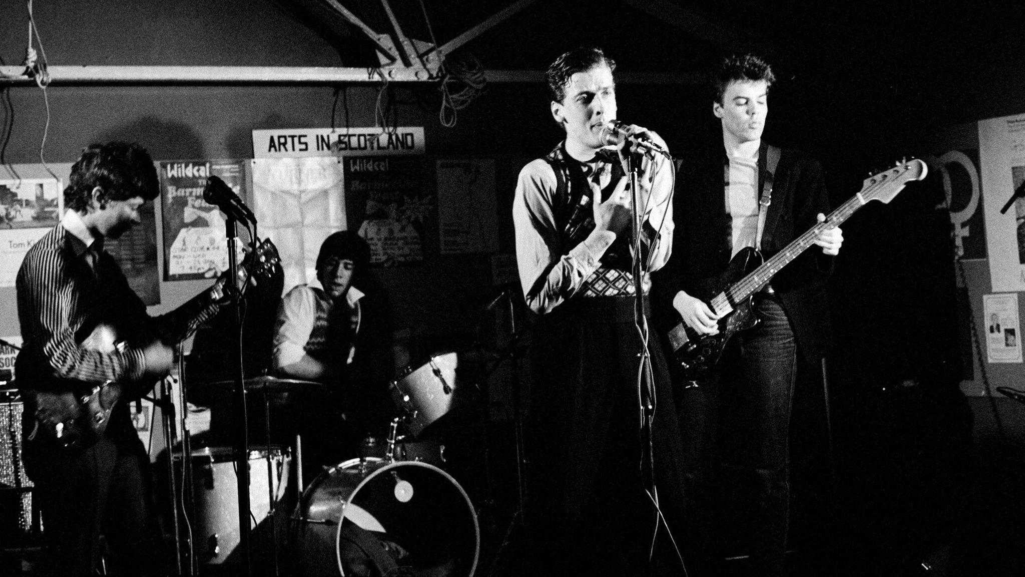 A black and white image of The Dreamboys performing on stage during the late 70s or early 80s. Peter Capaldi is singing at a microphone and there are two guitarists and a drummer