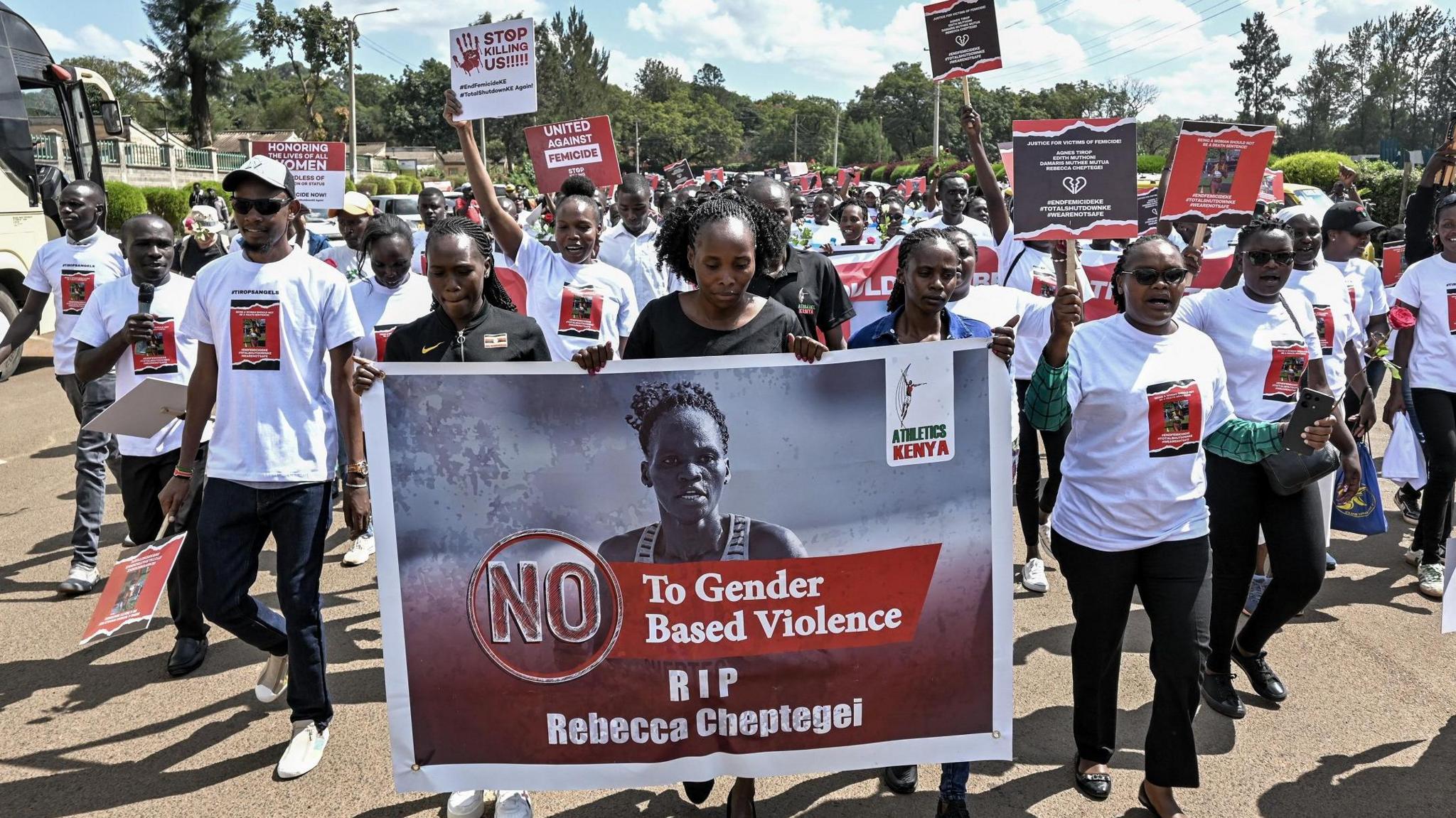 Protestors in white t-shirts walk down a street towards the camera, some waving signs while the three women at the front of the column hold up a large banner which includes a picture of Rebecca Cheptegei and reads "No to gender-based violence, RIP Rebecca Cheptegei"