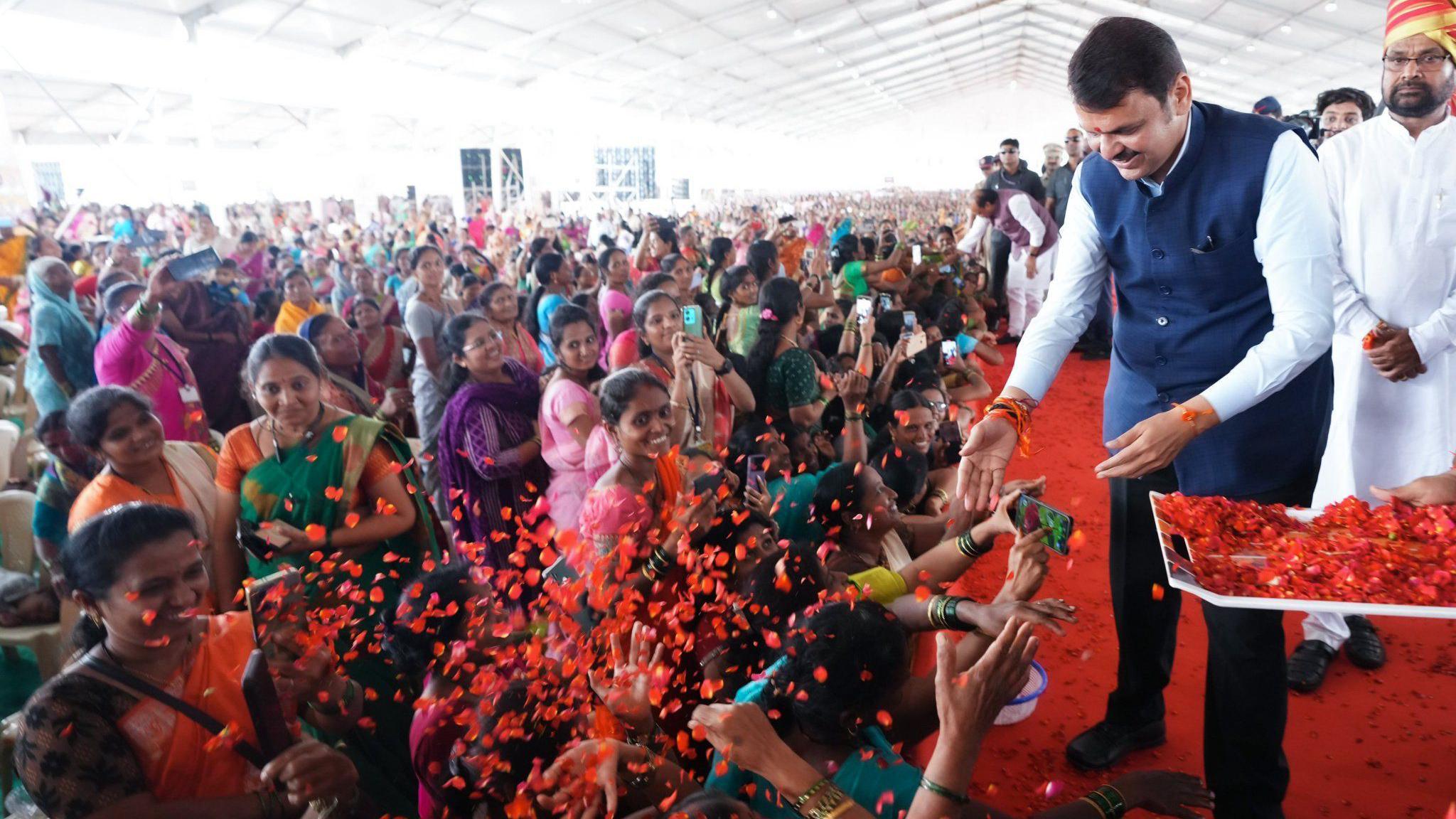Devendra Fadnavis dressed in a blue jacket, white shirt and black trousers, greets women, as BJP workers spray flower petals on the crowd 