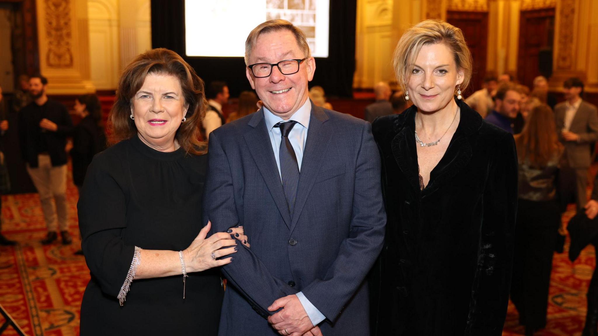 Two women and a man stand in a room full of people. On the left, Elaine C Smith, who has brown hair and is wearing a black dress with jewels on the sleeves. She is linking arms with Tommy Gormley. He is smiling, wearing glasses and a blue suit. On the right is Sarah Purser, who has blonde hair which is tied back and showing off some earrings. She is wearing a black jacket and a necklace.