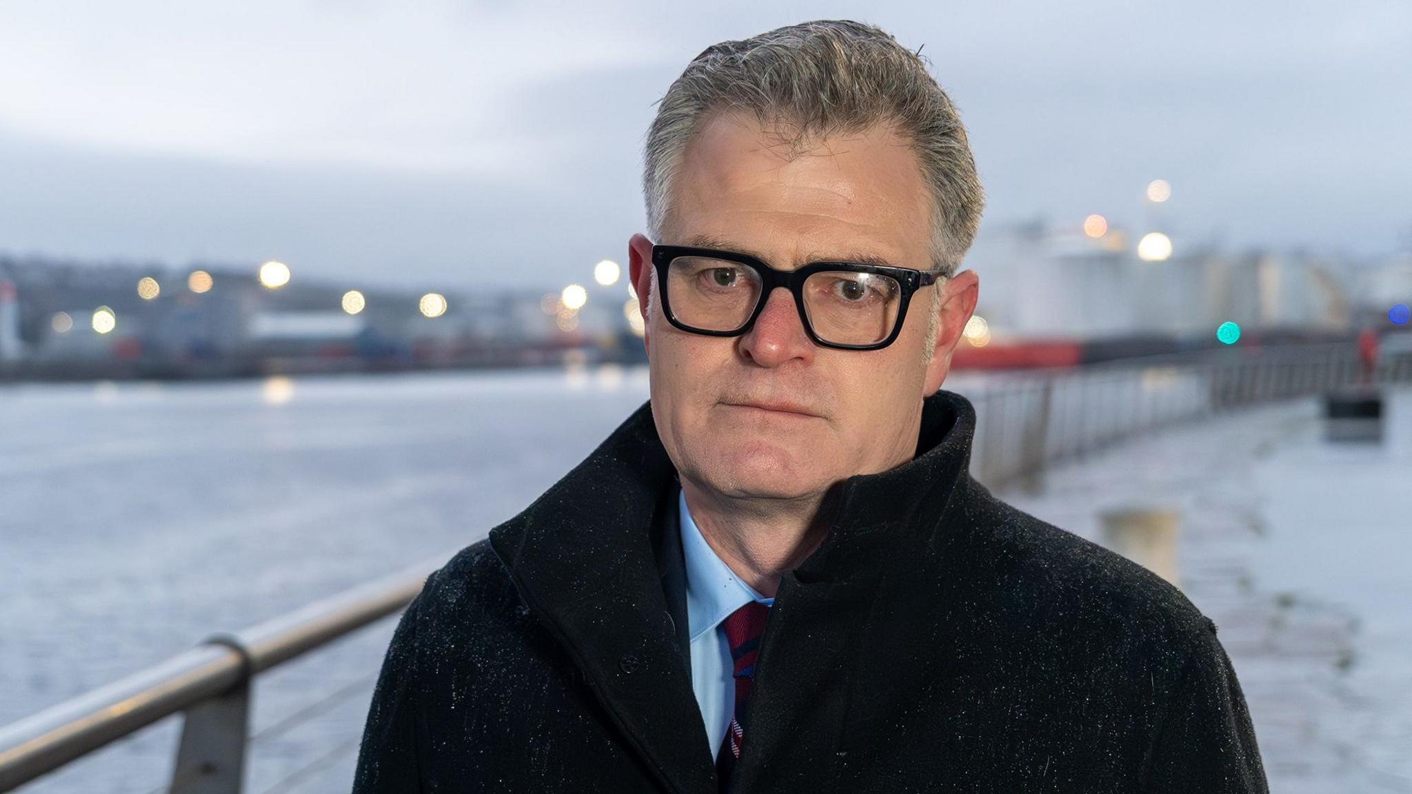 A man in glasses, wearing a dark jacket standing next to a barrier beside water