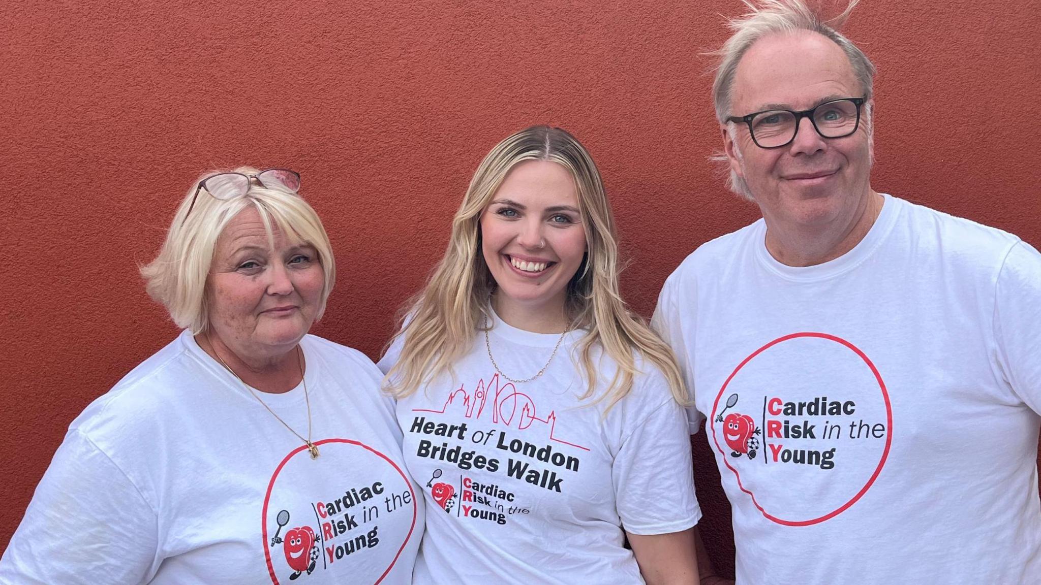 Two women and a man are stood together in front of a red wall. They are all smiling and wearing white T-shirts that display a Cardiac Risk in the Young logo. The first woman, on the left of the picture, has short blonde hair and a pair of glasses on top of her head, the second woman, in the centre of the picture has long blonde hair and a T-shirt that also displays the words: Heart of London Bridges Walk. The man on the right of the picture has short grey hair and is wearing glasses.