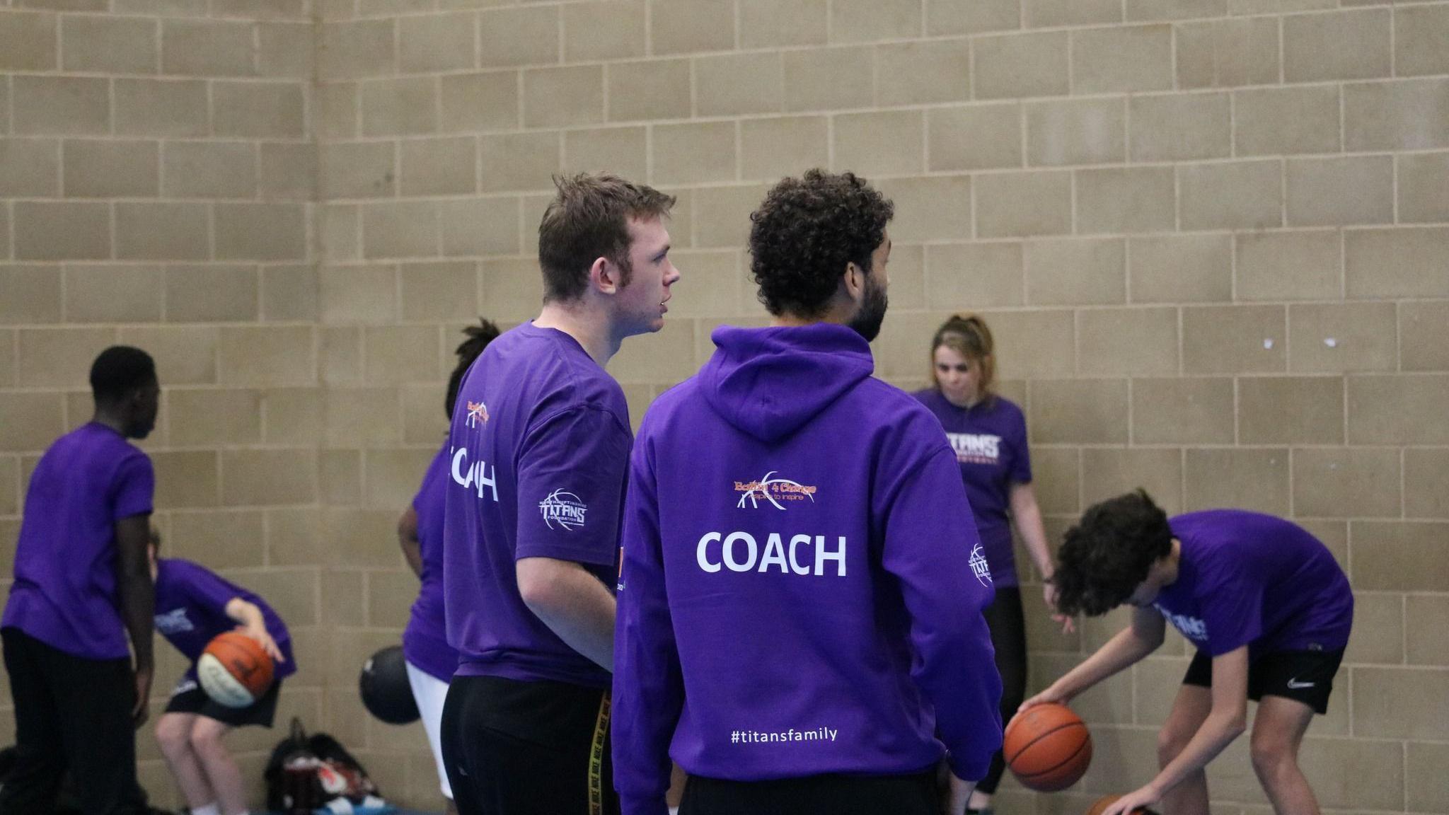 Several people in purple hoodies and t-shirts playing basketball next to a grey brick wall. 