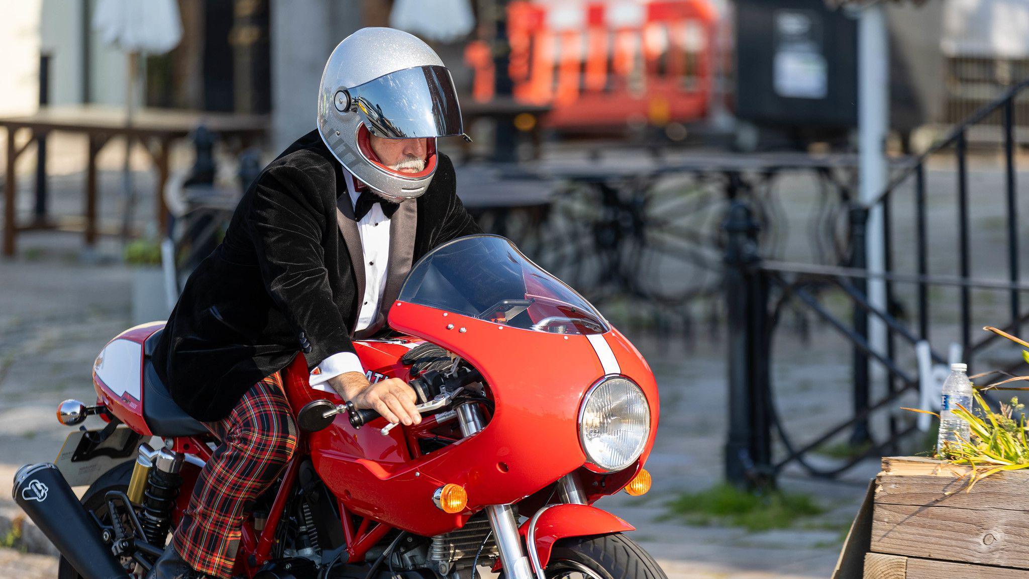 A man in a black tuxedo jacket and bow tie with red tartan trousers riding a red motorcycle.