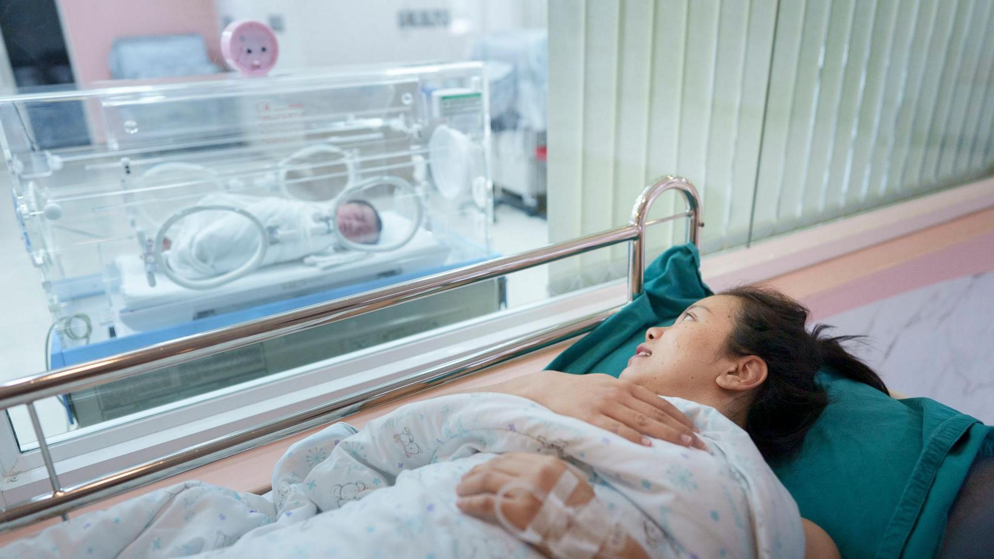 Mother in hospital lying on a bed with a baby in an incubator next to her.