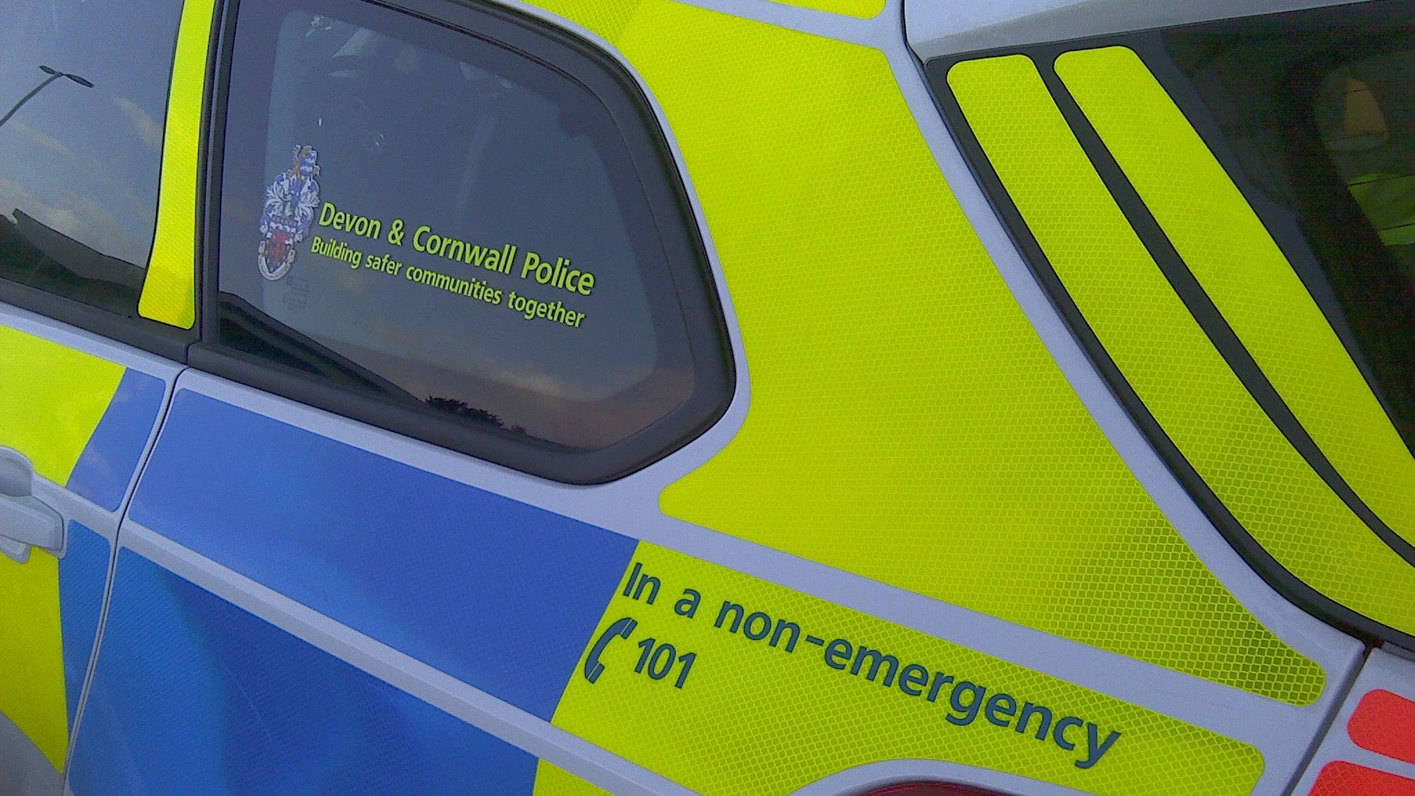 The rear end side of a police car with the Devon and Cornwall Police branding on the side. It has a non-emergency number on the side also. 