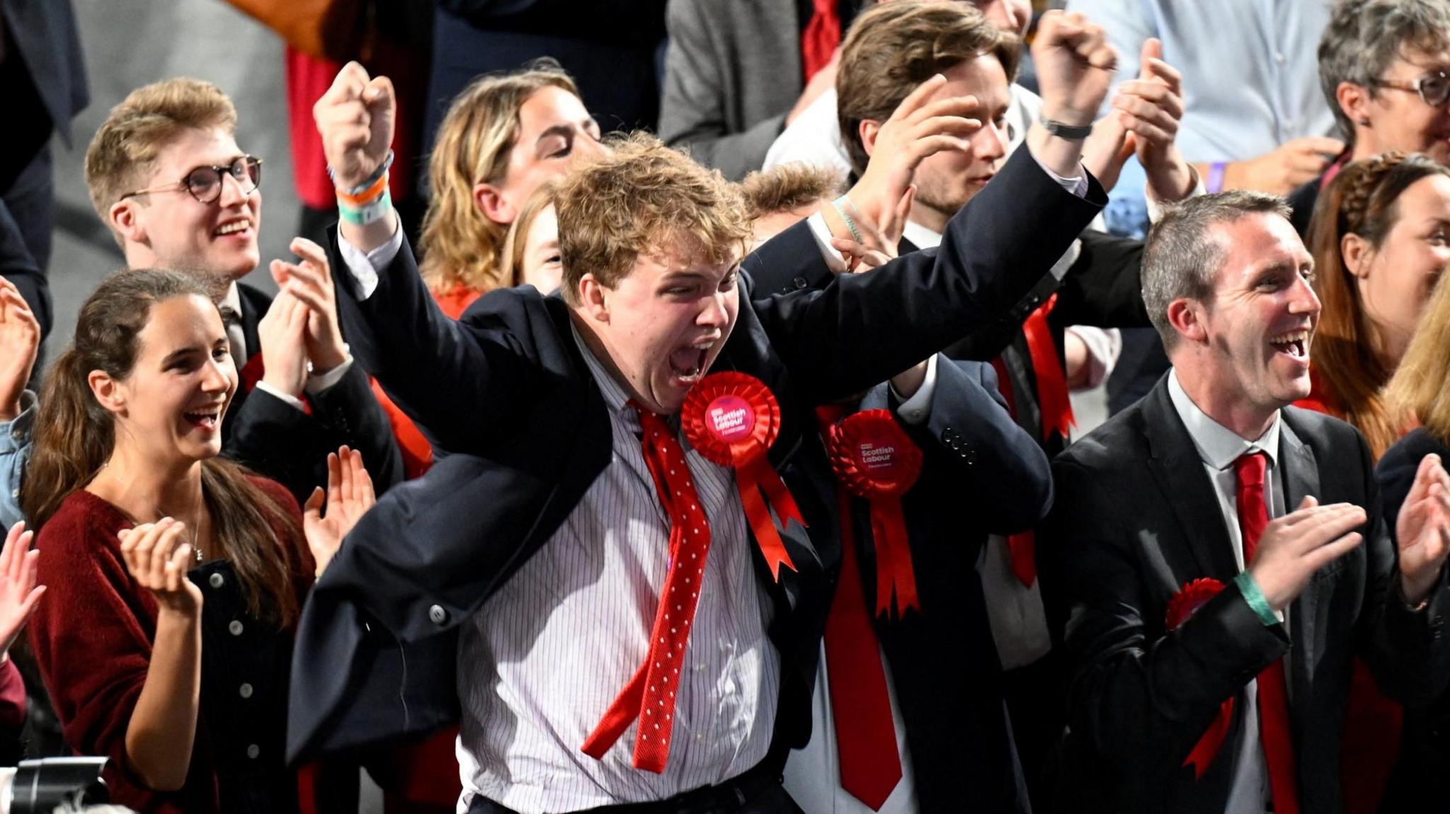 A group of Labour supporters smile and applaud at a counting centre in Glasgow. A man in the centre is wearing a dark suit with a red tie and a red rosette. He has his arms raised and is shouting in celebration.