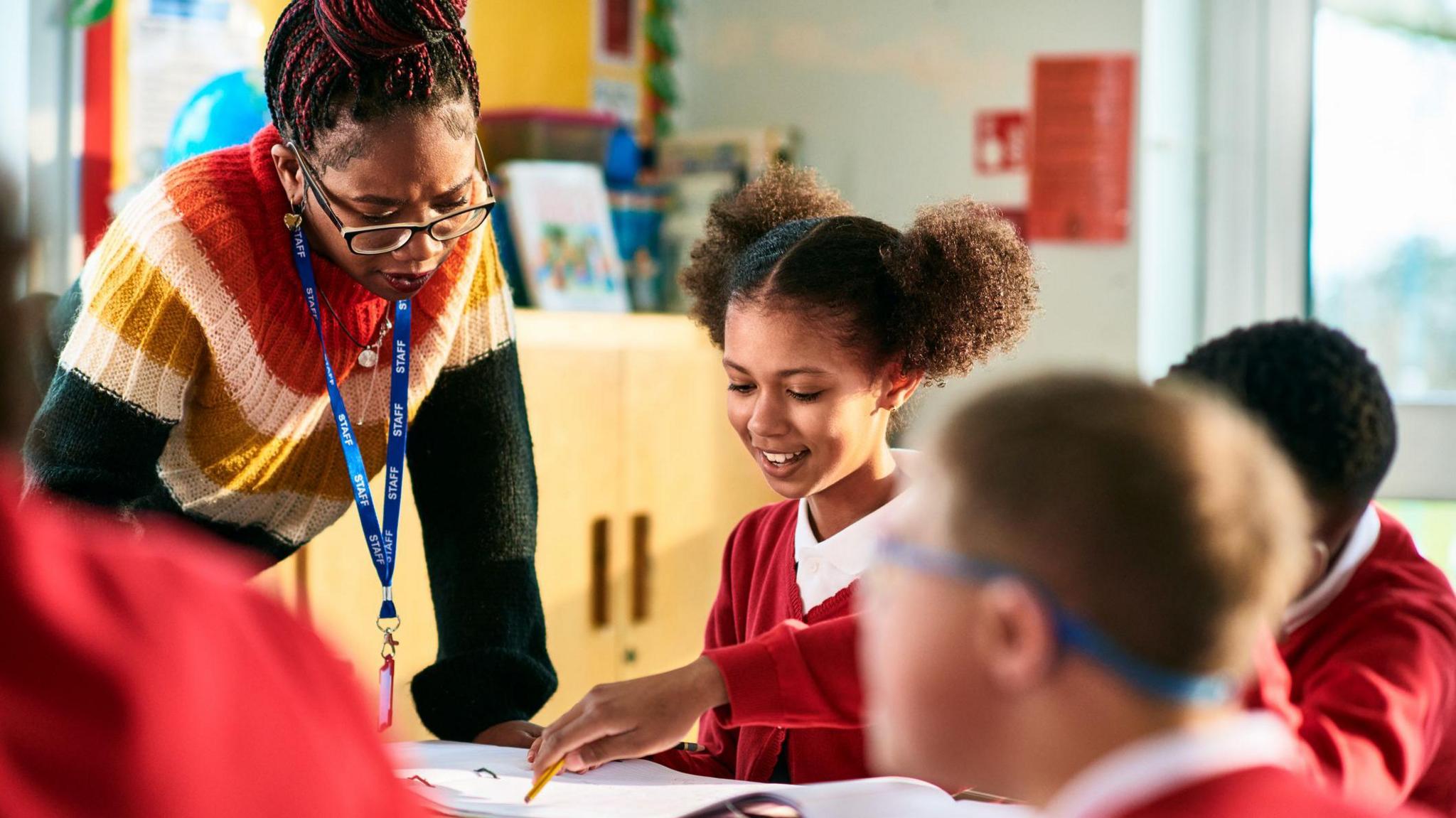 A teacher wearing a brightly coloured knitted jumper leans over a table in a classroom where children in red uniform cardigans are doing their work. In focus is a girl who is smiling down at the paper in front of her.