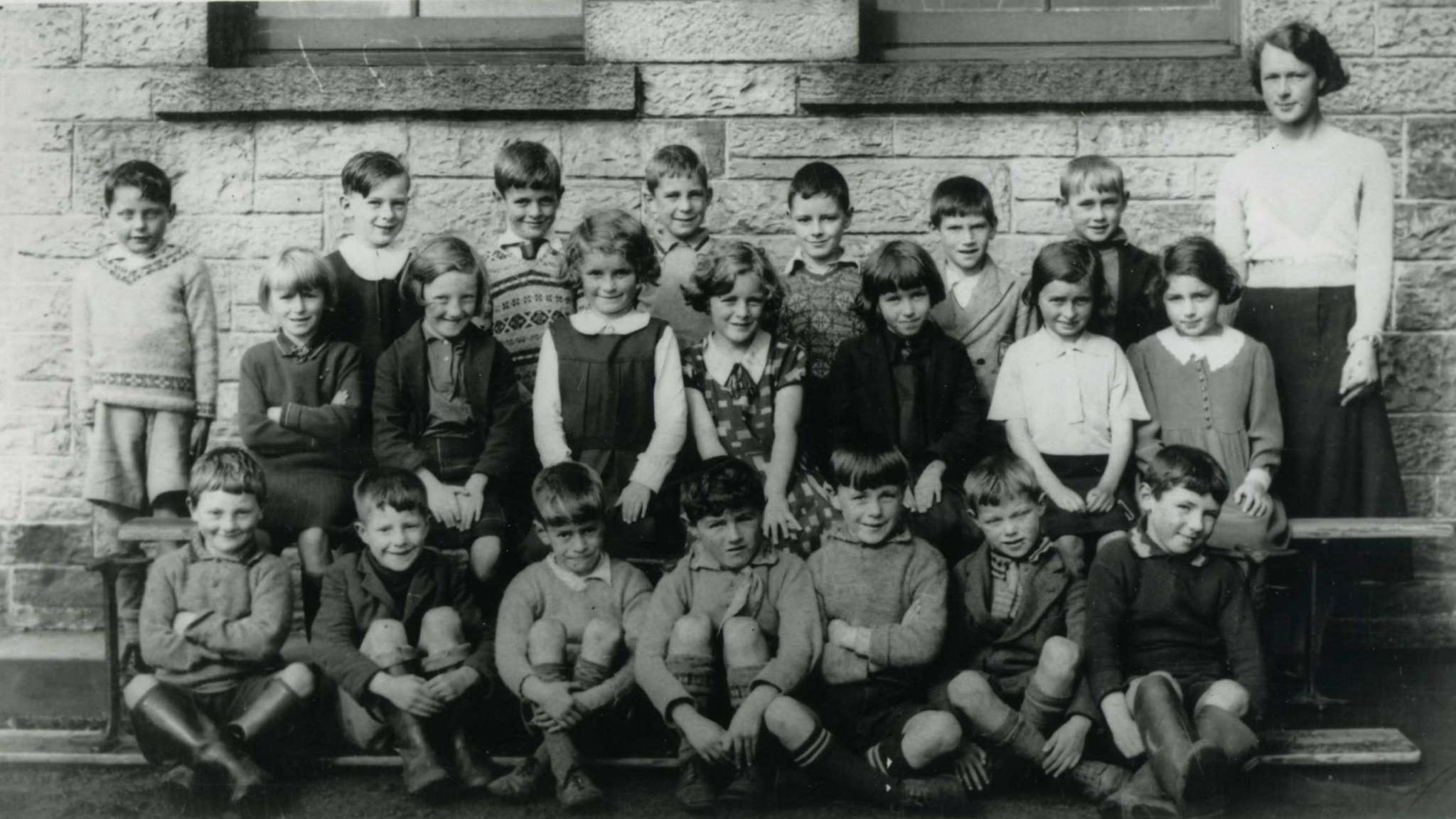 A black and white school photo from the 1930s. Three rows of 22 children. Robert Shaw is standing slightly apart from his classmates on the top left, wearing shorts and a light v-neck jumper with a pattern around the neck. A female teacher in a light top and dark skirt is standing at the other end of the rows. They are against a stone wall.