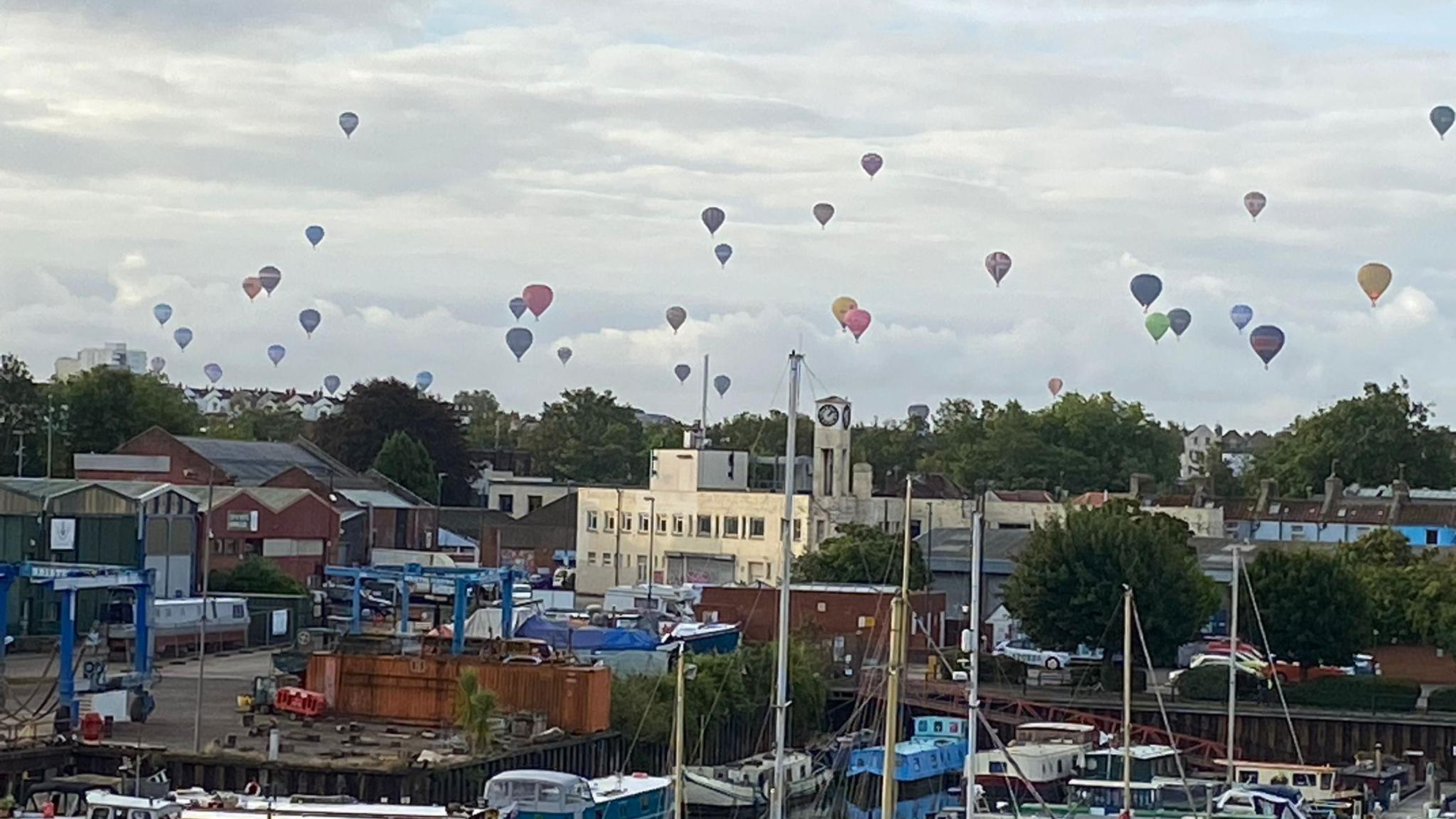 Balloons flying over a harbour with many boats in the water.