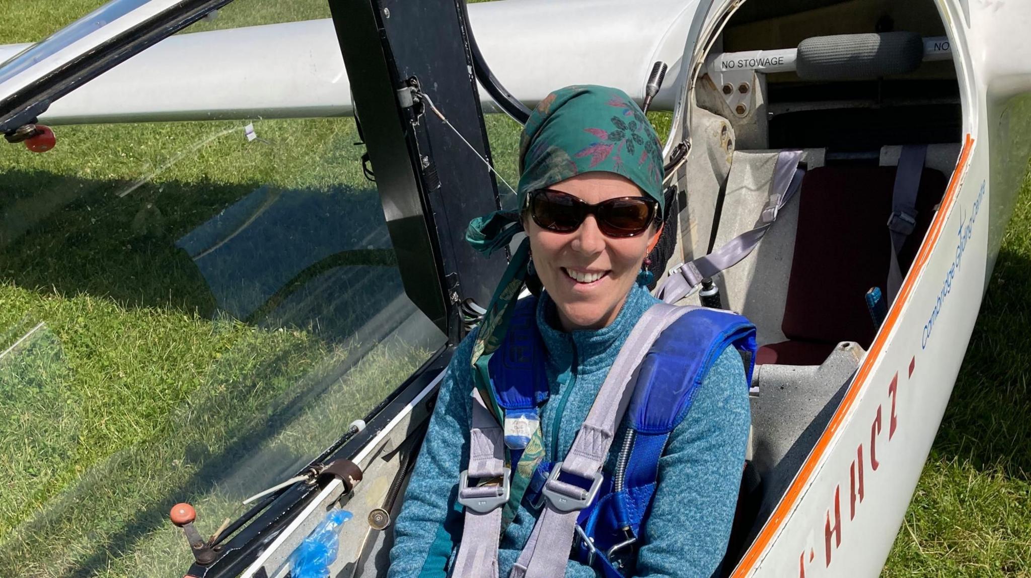 Julia Beeden sits in the cockpit of a glider. She has a grey seatbelt on. Julia has a green scarf around her head where she has lost her hair because of chemotherapy and is wearing a green, zip up top. She is smiling and has sunglasses on.