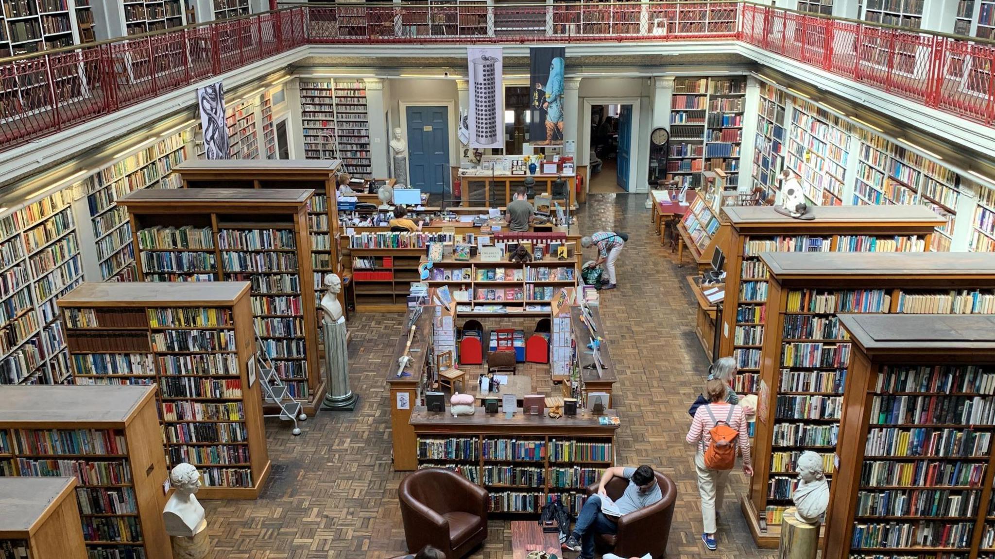 A large room filled with shelves of books and a walk way going down the centre. People browse books in the large, lit room.