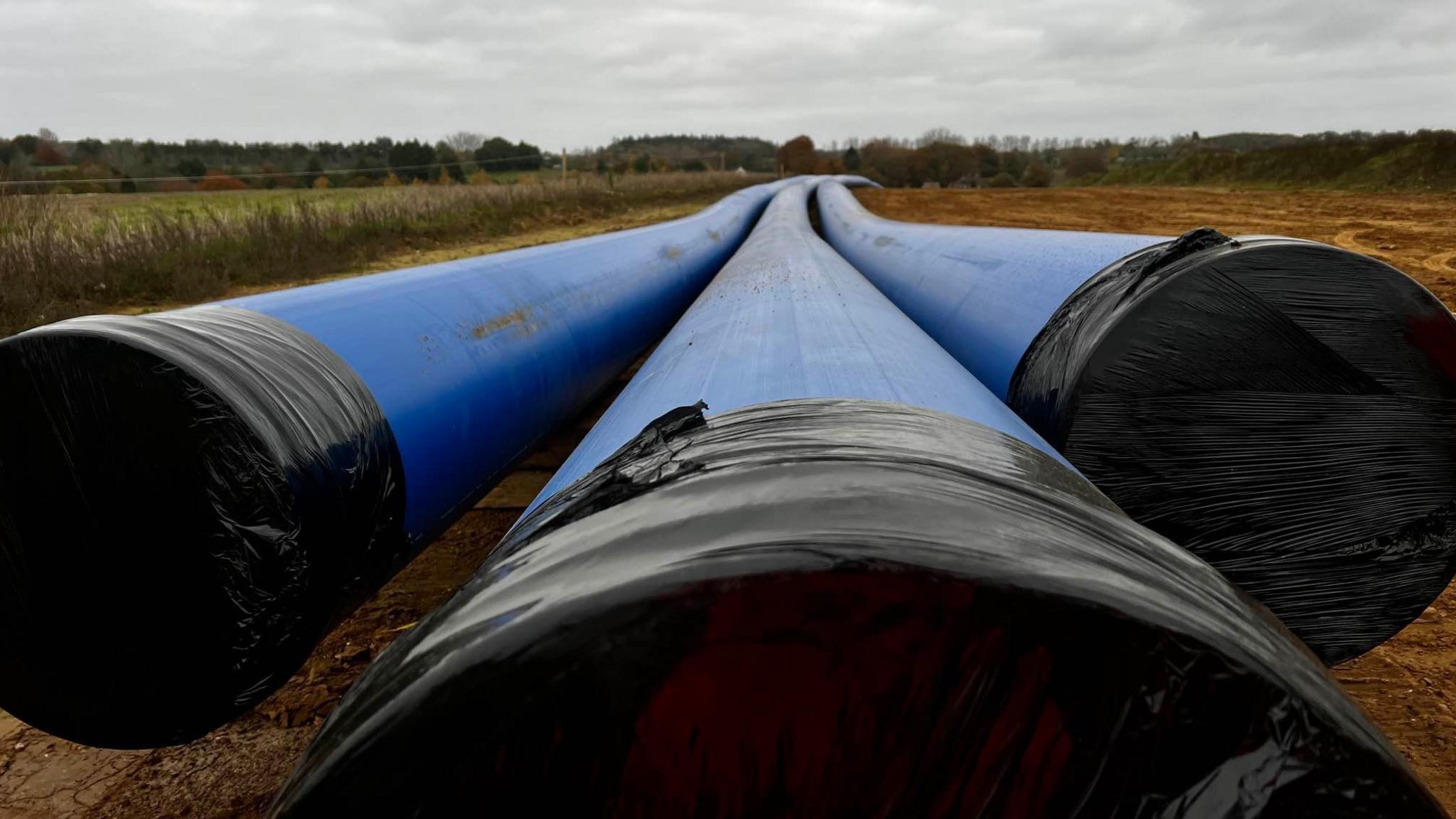 A view of three large blue water pipes that span off into the distance. They sit in a field and their openings have been covered by black sheeting. Grass and fields can be seen either side of the pipes